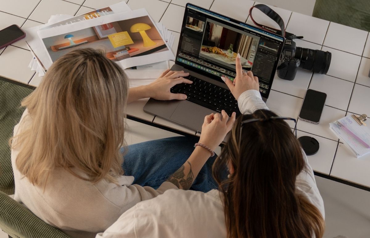 Zwei Personen schauen gemeinsam auf einen Laptop auf einem weißen Fliesentisch. Die rechte Person zeigt auf den Bildschirm mit Bildbearbeitungssoftware (Speisen und Getränke), die linke tippt. Kamera, Smartphone und Dokumente liegen auf dem Tisch.