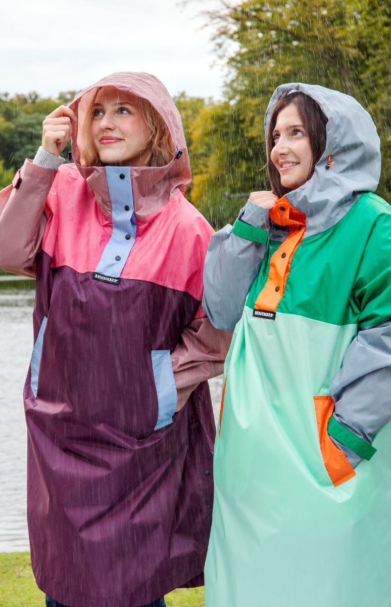 Zwei lächelnde Frauen in bunten Kapuzen-Regenponchos stehen im Regen. Links trägt eine einen rosa-violetten Poncho, rechts eine grün-mintfarbenen. Beide blicken froh in den Himmel, während Tropfen fallen. Im Hintergrund: Bäume und Gewässer.