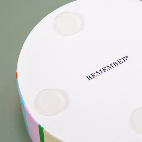 The underside of a round, colourful coaster: a white surface featuring a diagonal black ‘REMEMBER®’ logo and three transparent, round anti-slip pads. The edge features vertical stripes in pastel and bold shades.