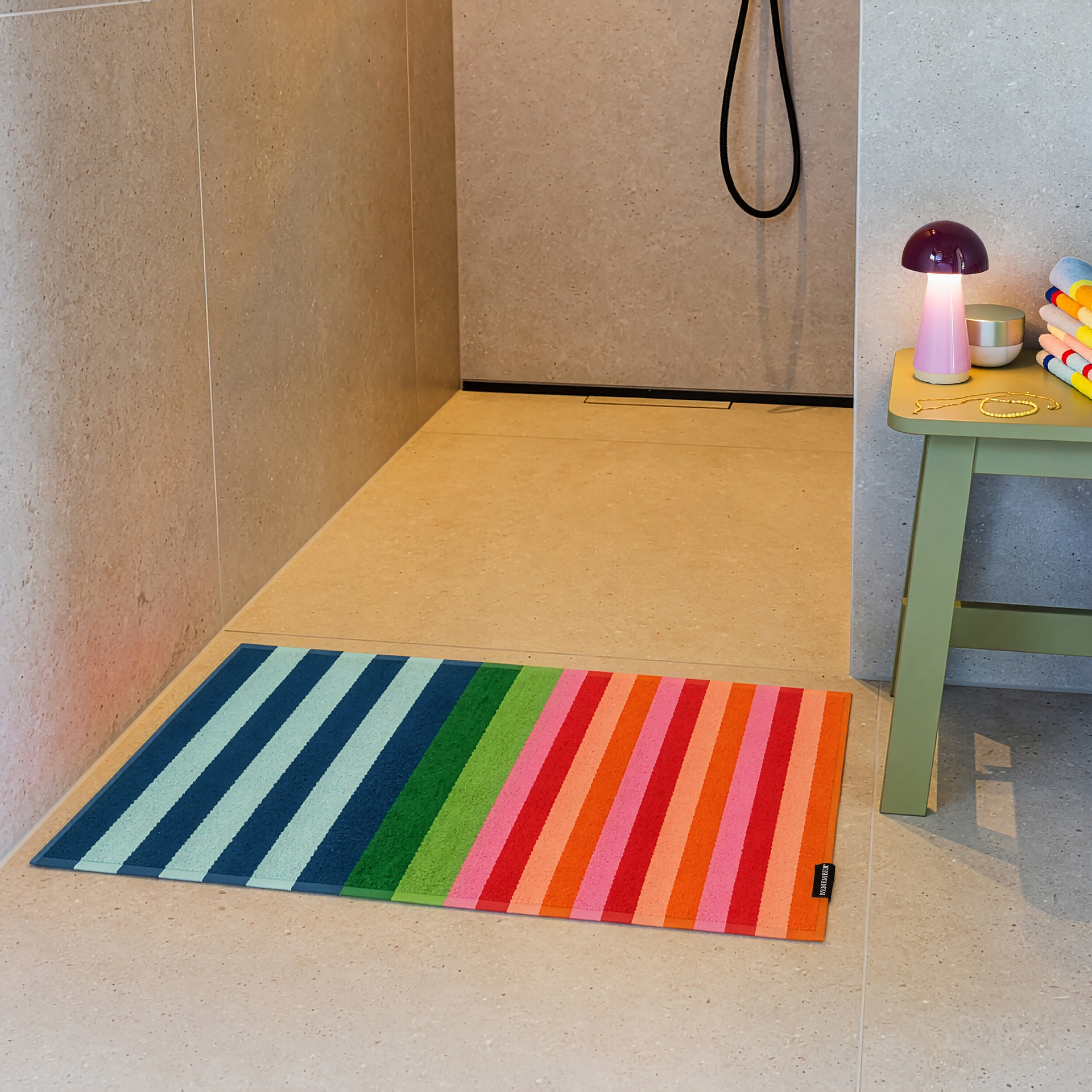 Un tappeto da bagno colorato con strisce diagonali di vari colori giace sulle piastrelle chiare di un bagno moderno. Le strisce vanno dal blu al verde, al rosa, al rosso e all'arancione. A destra uno sgabello con lampada rosa e asciugamani.