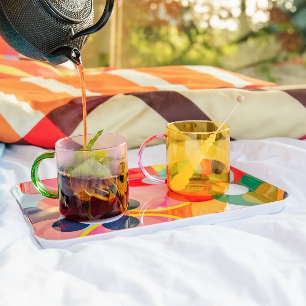 Une théière noire verse du thé dans une tasse en verre à menthe avec anse verte. À côté, une tasse en verre jaune avec un bâtonnet de sucre candi repose sur un plateau coloré. Le tout sur un drap blanc, fond ensoleillé.