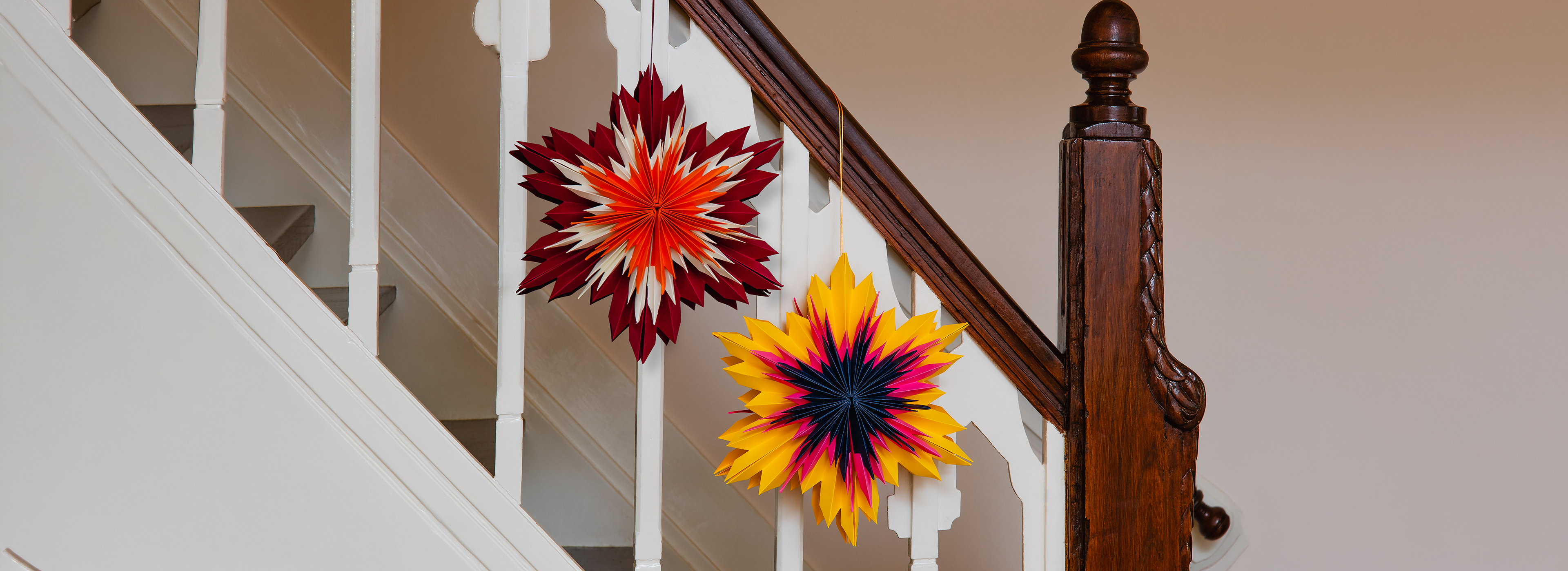 Zwei bunte, gefaltete Papiersterne hängen an einer Treppe mit weißem Geländer und dunklem Holzhandlauf. Der obere ist rot, orange und weiß, der untere gelb, pink und dunkelblau – beide geben dem Treppenhaus eine festliche Note.