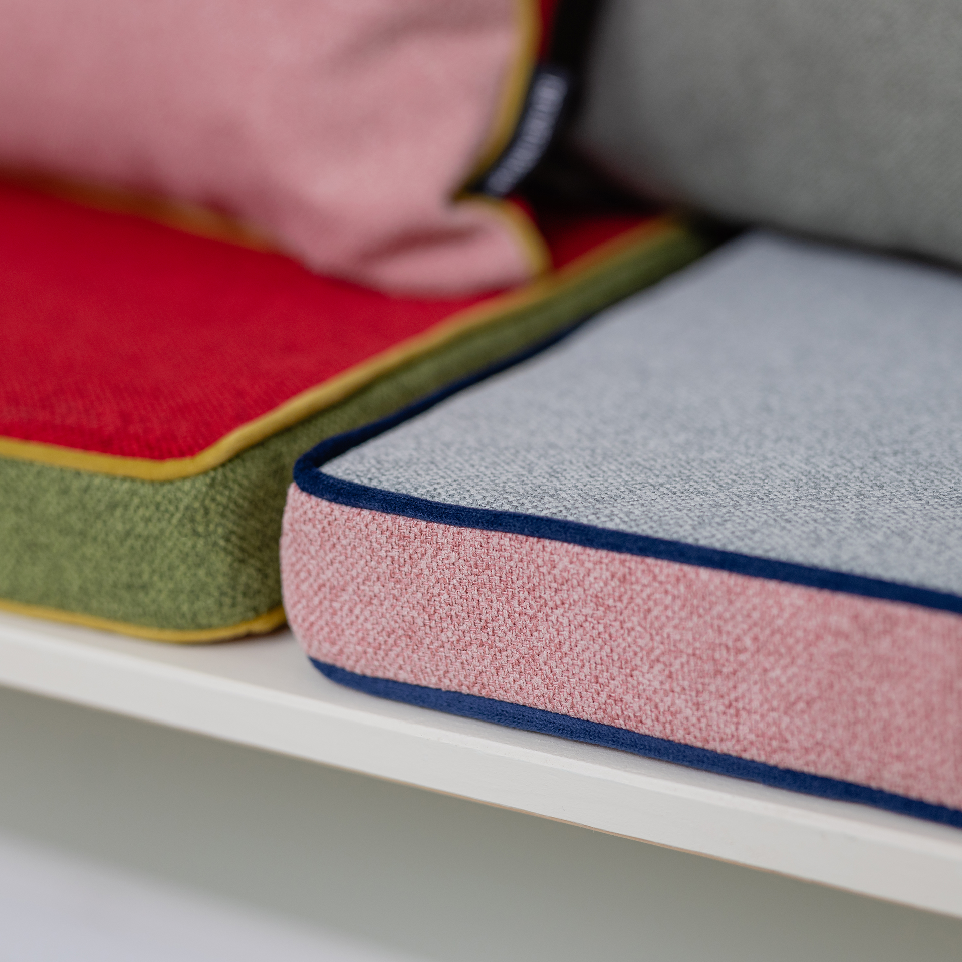 Detailopname van gekleurde zitkussens met biezen op een witte plank. Het voorste kussen is grijs met een roze zijkant en blauwe rand. Daarachter ligt een rood, groen en geel kussen.