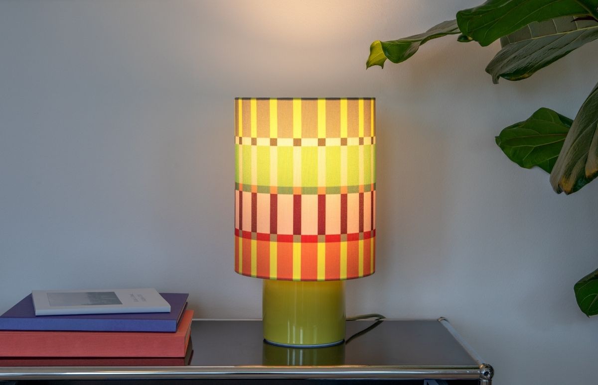 Helle Tischlampe mit gelbem Sockel auf dunkler, reflektierender Oberfläche. Zylindrischer Schirm mit bunten Streifen und Quadraten in Gelb, Grün, Orange, Rot und Braun, warm leuchtend. Links Bücherstapel, oben rechts Zimmerpflanze.