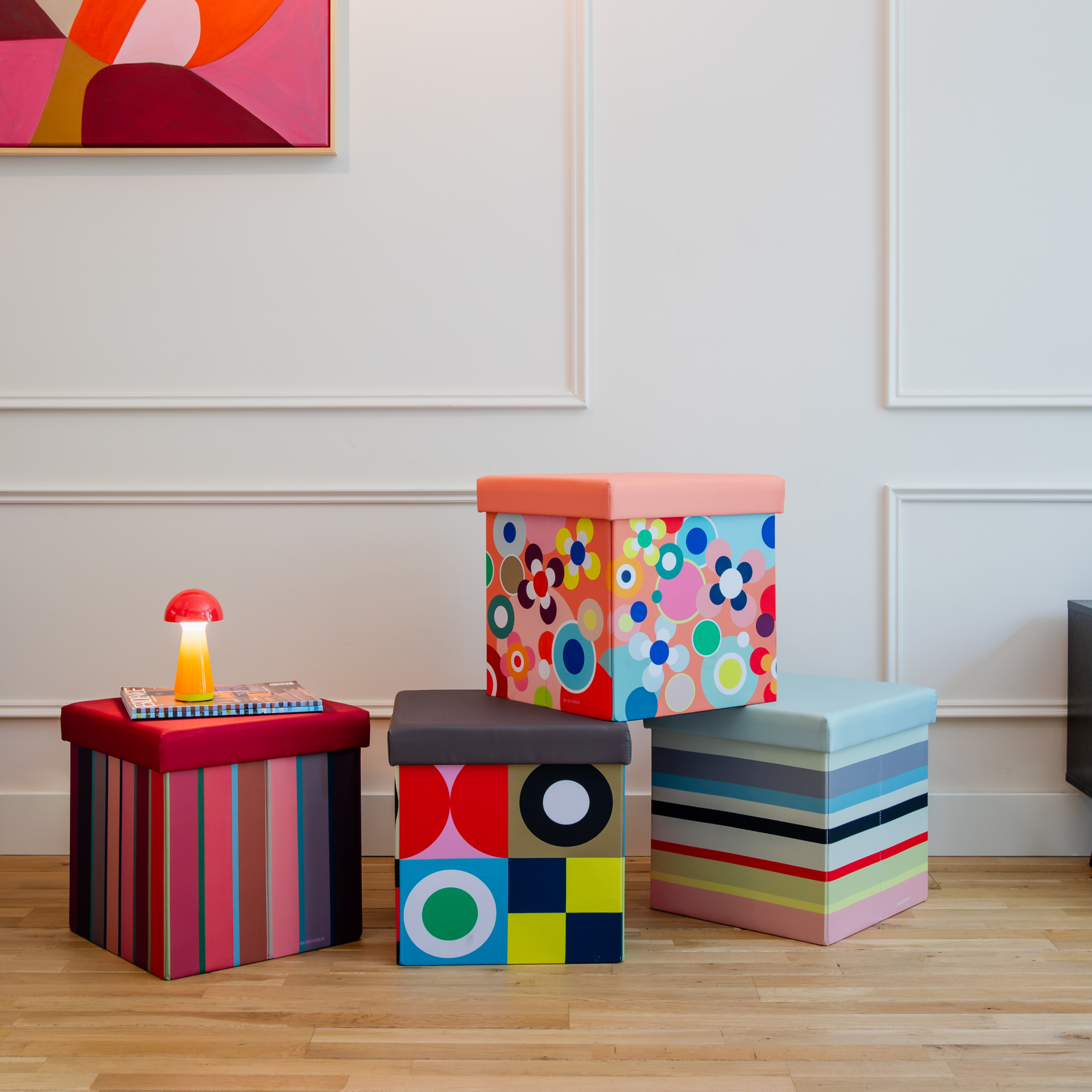 Vier farbenfrohe Sitzboxen mit Mustern wie Streifen, geometrischen Formen und Blumen stehen auf hellem Holzfußboden. Eine Pilzleuchte steht auf roter Sitzfläche. Die dekorativen Boxen bieten Stauraum und Sitzgelegenheiten.