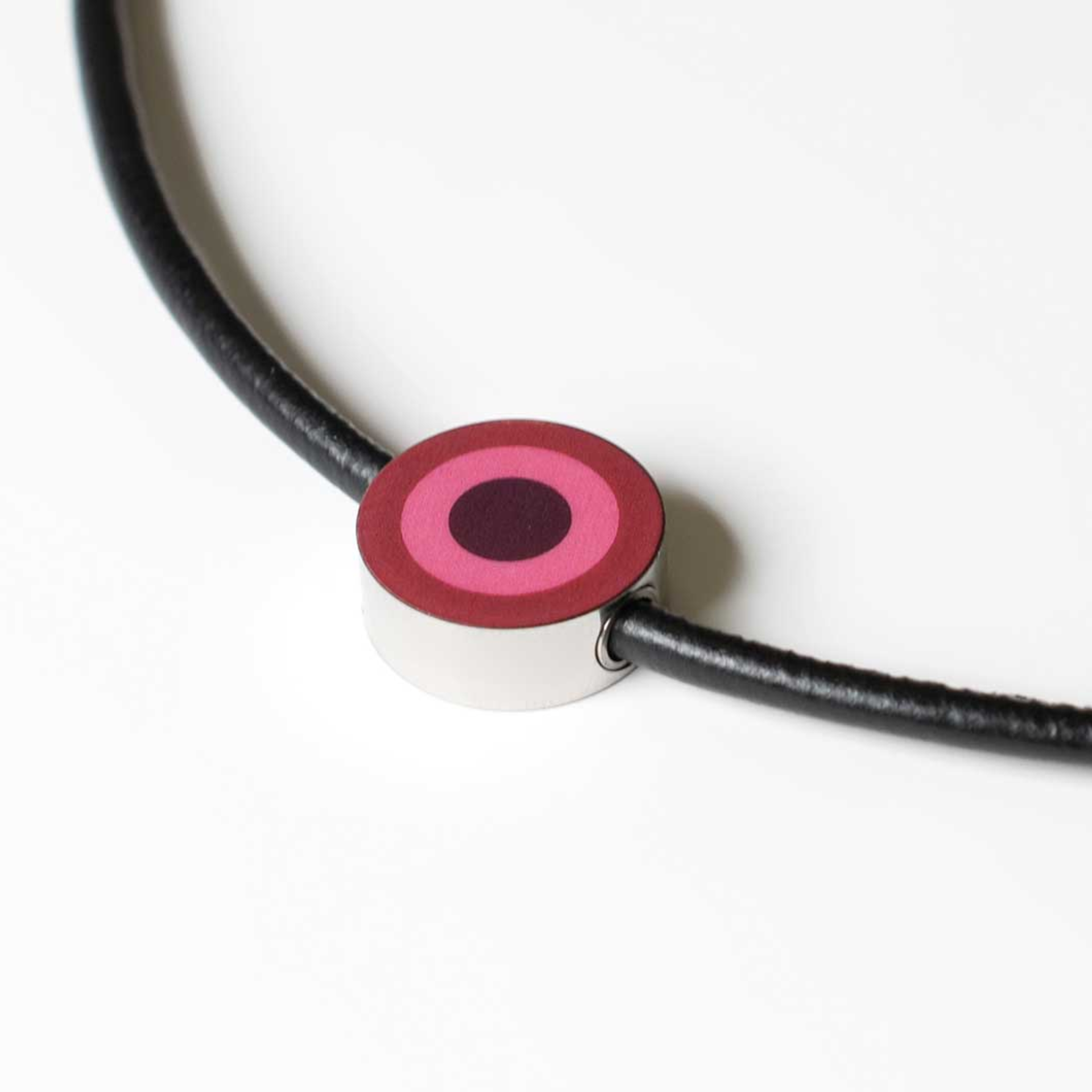 Pendentif bijou : Bijou rond avec des cercles concentriques en rose et rouge foncé sur un ruban noir. Le pendentif est argenté brillant.