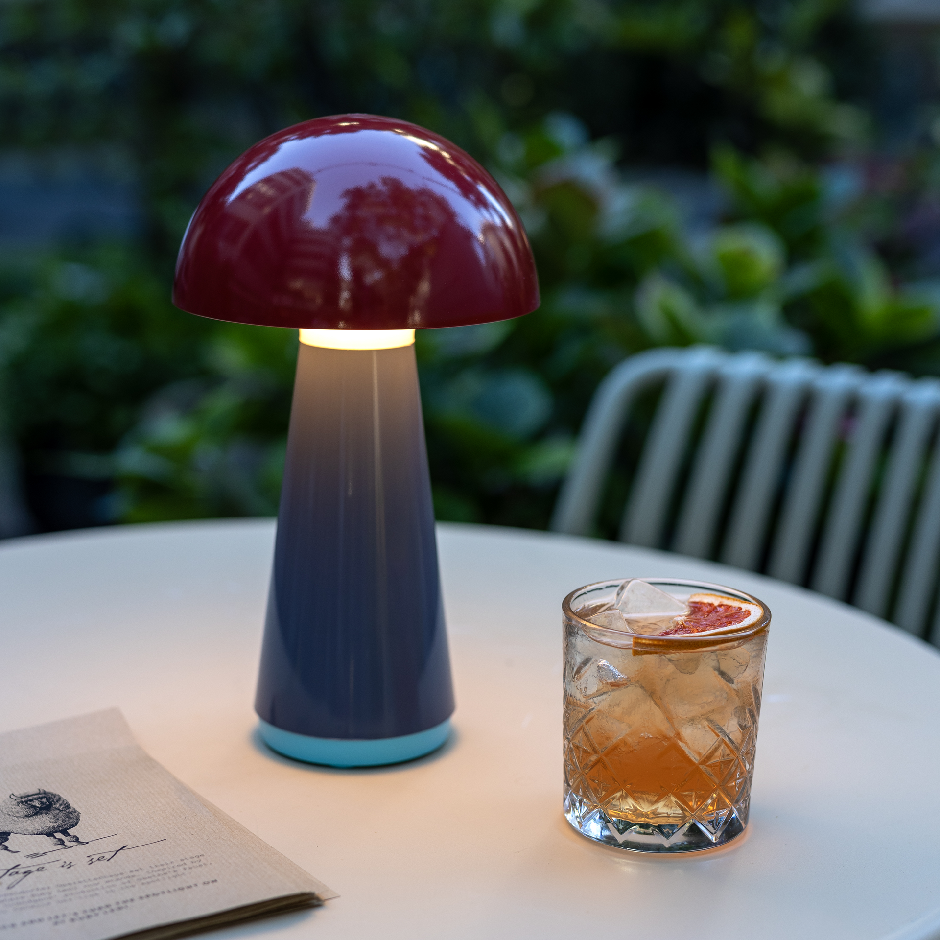 Een rode 'Bob' tafellamp in paddenstoelmotief schijnt naast een cocktailglas met ijsblokjes op een buitentafel. Op de achtergrond zijn groene planten zichtbaar.
