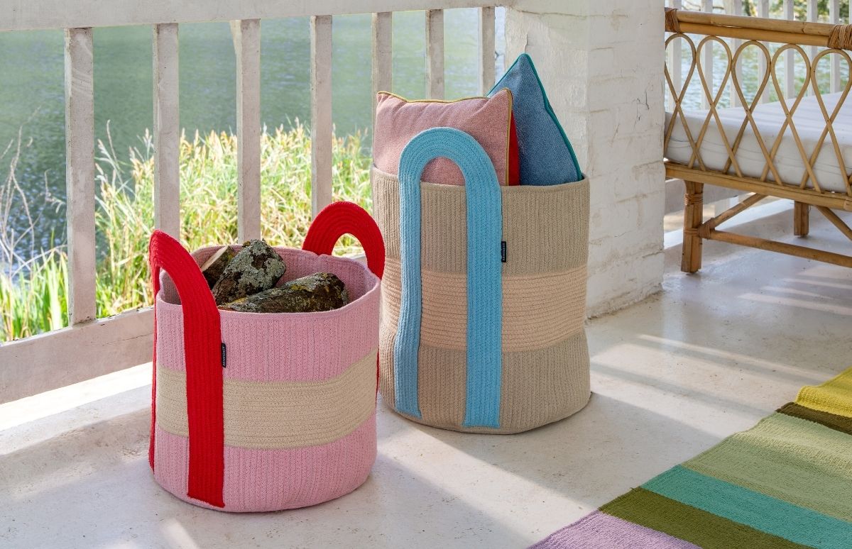 Zwei gestrickte Körbe stehen auf einer hellen Veranda. Der rosafarbene Korb mit roten Griffen enthält Holzscheite, der beigefarbene mit blauen Griffen ist mit rosa und blauen Kissen gefüllt. Im Hintergrund: Geländer, Schilf.