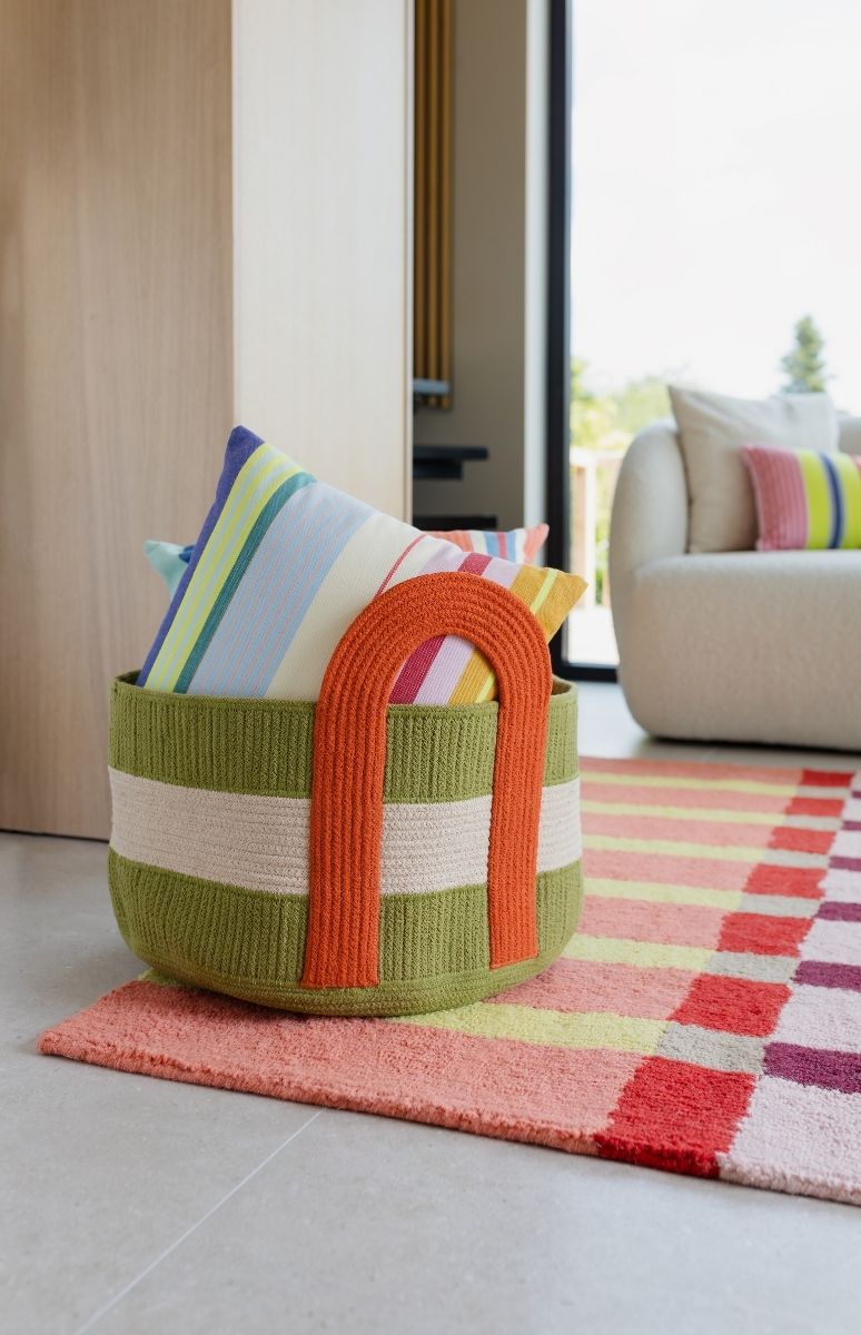 Ein grüner Strickkorb mit beigen Streifen und orangem Henkel steht auf einem bunten Teppich. Darin liegen mehrere gestreifte Kissen in Pastell- und Leuchtfarben. Im Hintergrund sieht man eine helle Couch und ein Fenster ins Grüne.