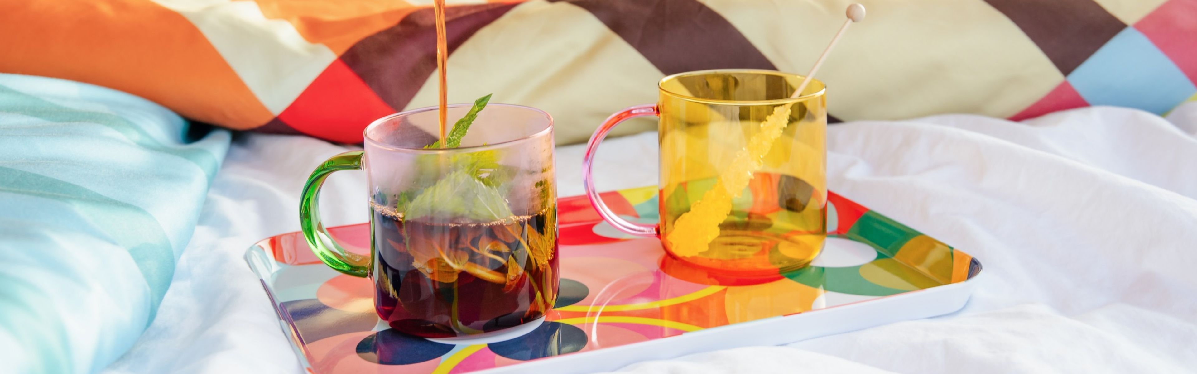 Deux tasses en verre colorées sur un plateau à motifs, sur un lit blanc. À gauche, du thé noir est versé dans une tasse à anse verte avec menthe. À droite, une tasse jaune à anse rose avec sucre candi.