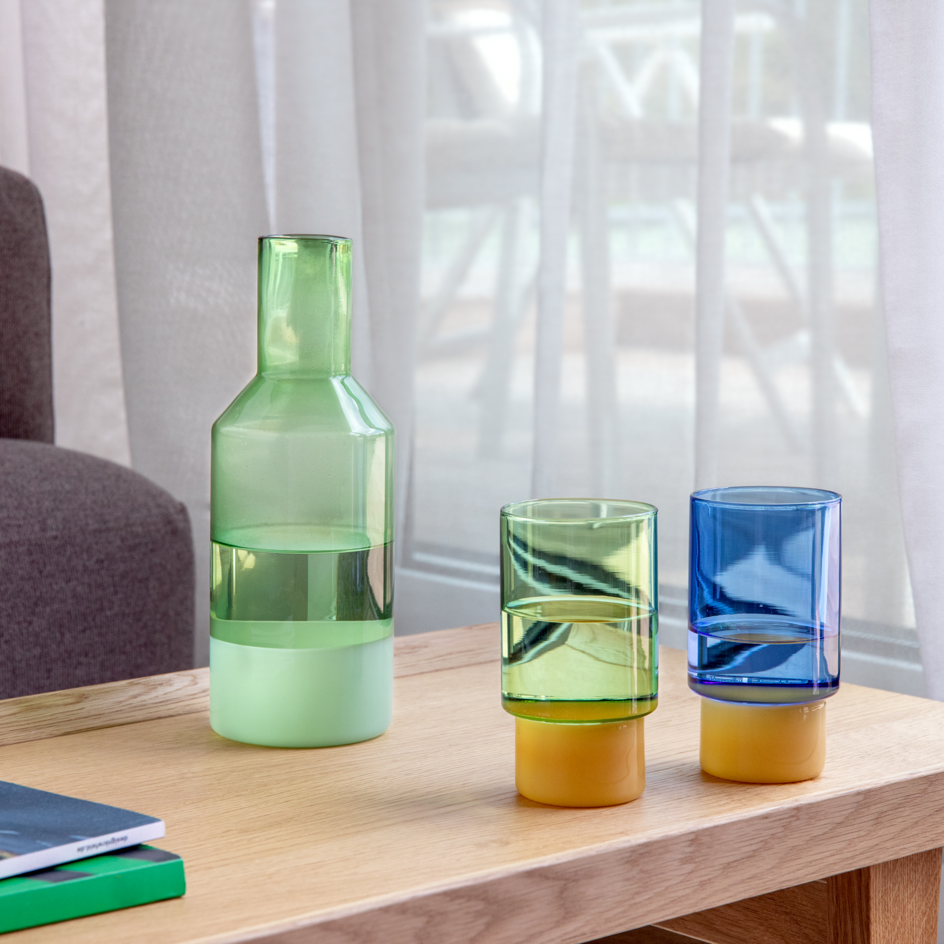 Drei farbige Borosilikatglas-Gefäße auf hellem Holztisch: links grüne Karaffe mit mattem, hellem Sockel. Daneben zwei Jill-Gläser, grün und blau getönt, beide mit gelbem Sockel. Teilweise mit Wasser. Hintergrund: weiße Vorhänge, Sessel.