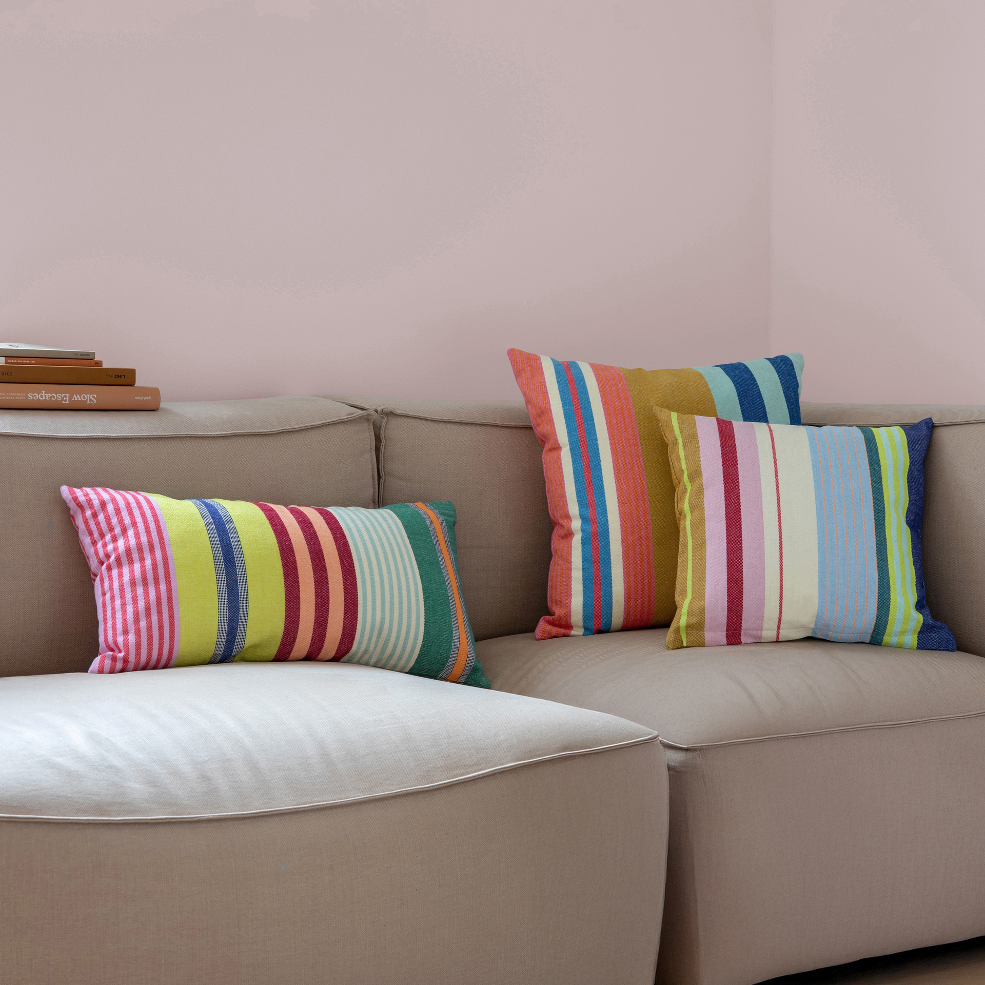 Close-up: drie 'Belize' katoenen kussens met kleurrijke strepen liggen op een beige bank voor een roze muur.