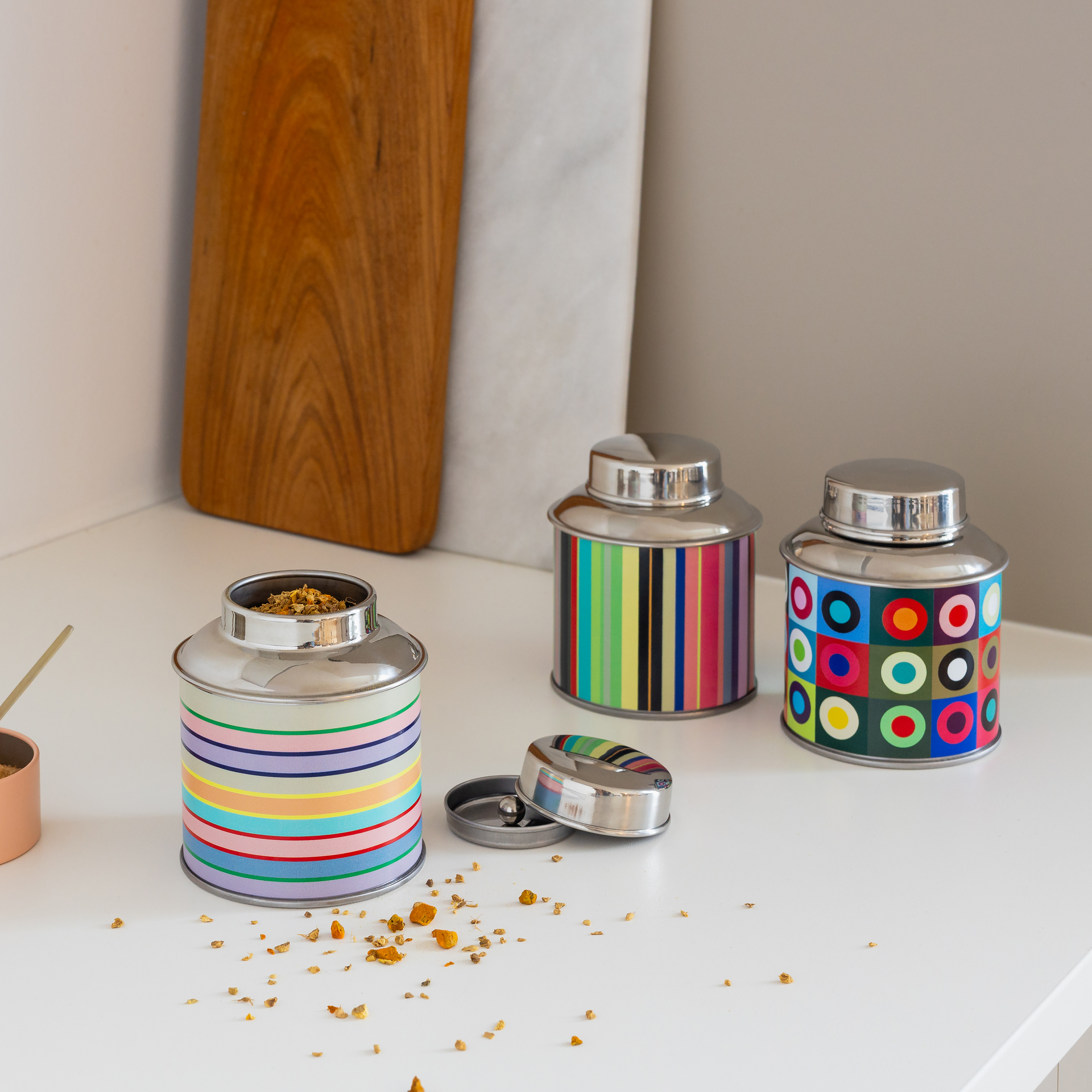 Round stainless steel tea caddy featuring a vibrant, multicoloured square pattern and colourful concentric circles. Topped with a shiny silver-coloured lid. Designed for storing tea; seals tightly to keep the contents fresh.