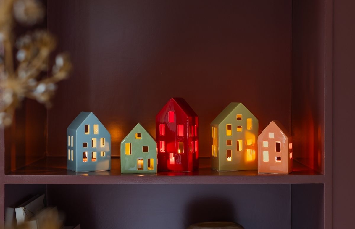 Fünf bunte Keramik-Lichthäuser stehen auf einem Holzregal vor dunkler Wand. Sie sind unterschiedlich hoch und in Hellblau, Hellgrün, Rot, Lindgrün und Hellorange. Warmes Licht strahlt durch Fenster und Türen.