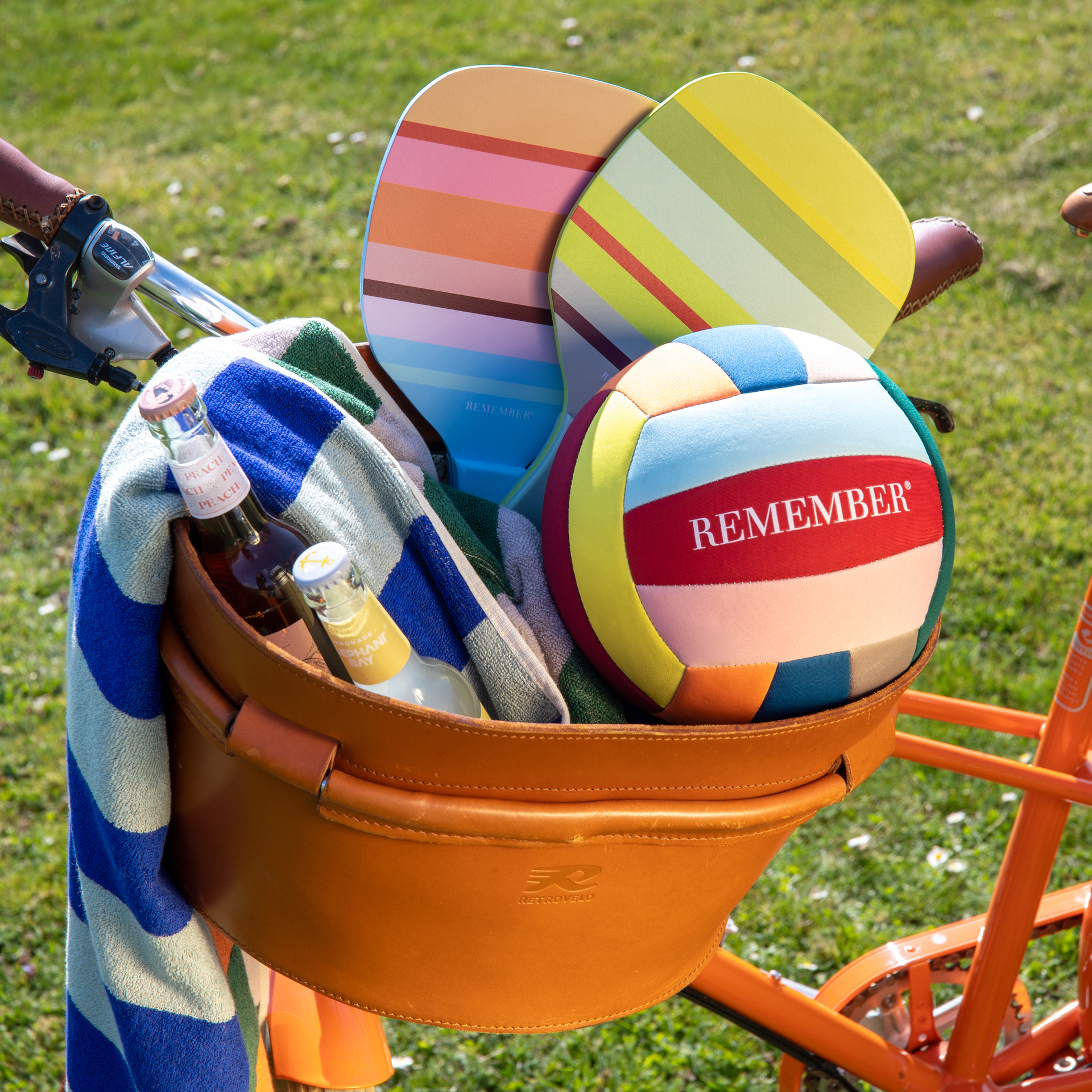 Bicycle basket with beach tennis set: racket, ball, drinks and towel in colourful stripes on grass.