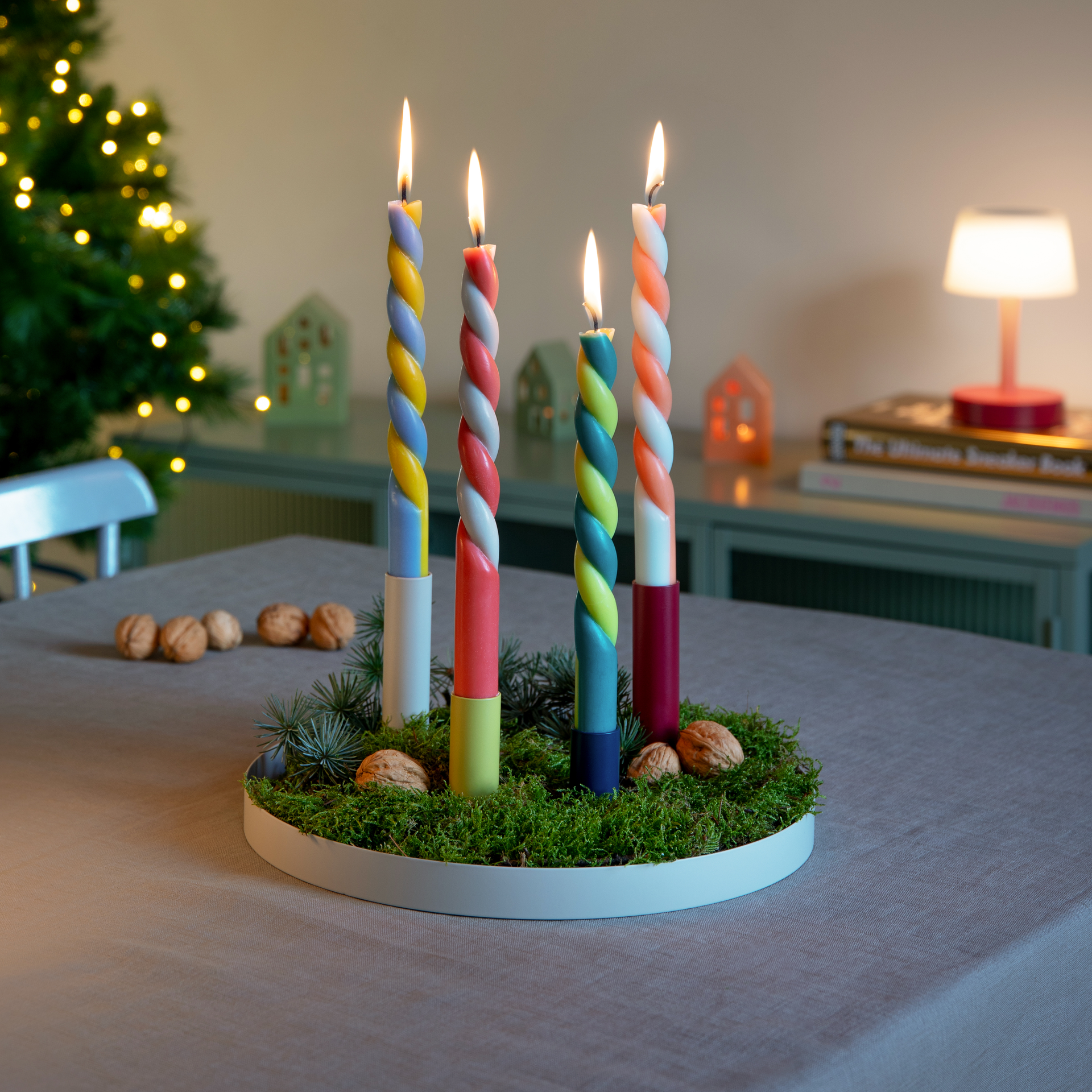 Portacandele magnetico "Luna" con quattro candele colorate e accese su un vassoio rotondo con muschio, noci e rami di abete, con un albero di Natale sullo sfondo.