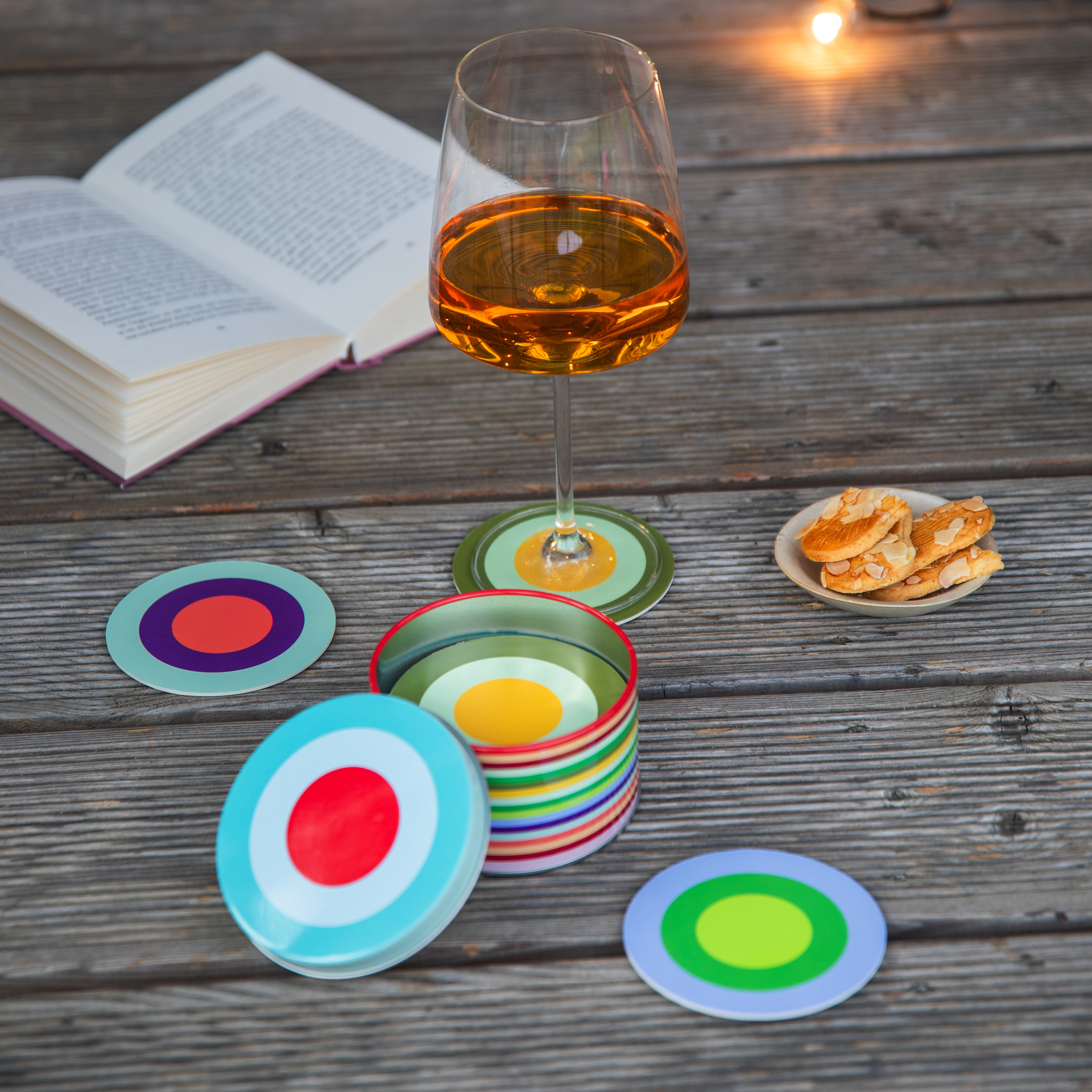 A table setting with the 'Ronda' coaster set, a glass of wine, biscuits and a book on a wooden surface. The coasters are colourful and circular.