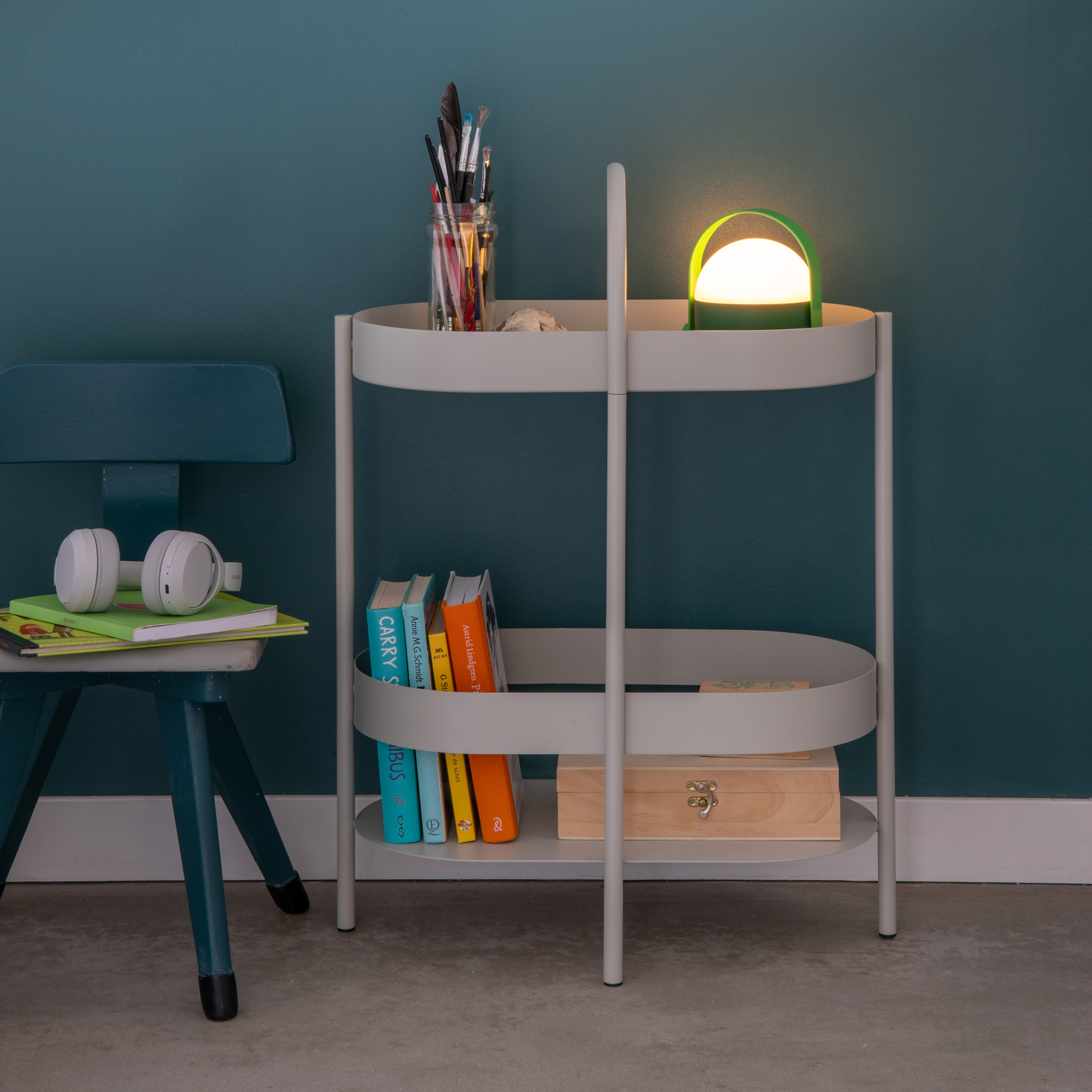 La consola auxiliar Barbara, de color claro, cuenta con dos estantes. En el estante superior hay lápices, lápices de colores y una lámpara verde. En el estante inferior hay libros y una caja de madera. Al fondo, una pared verde.