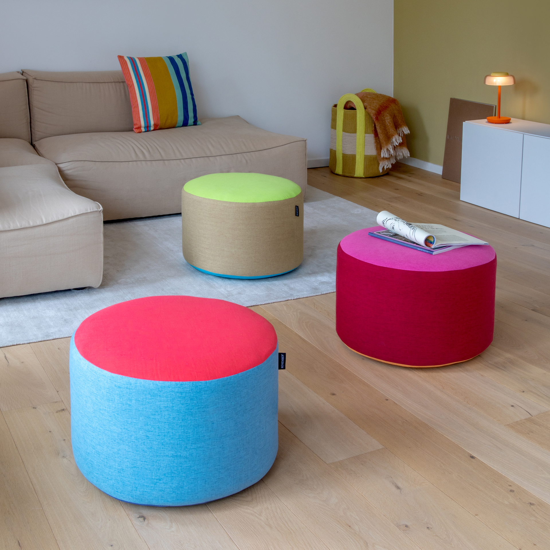 Três pufes redondos e coloridos na sala de estar. O da frente é azul-celeste com topo vermelho-coral; o do meio é bege com topo verde-neon e borda azul. Um pufe vermelho-escuro com topo rosa tem uma revista em cima.