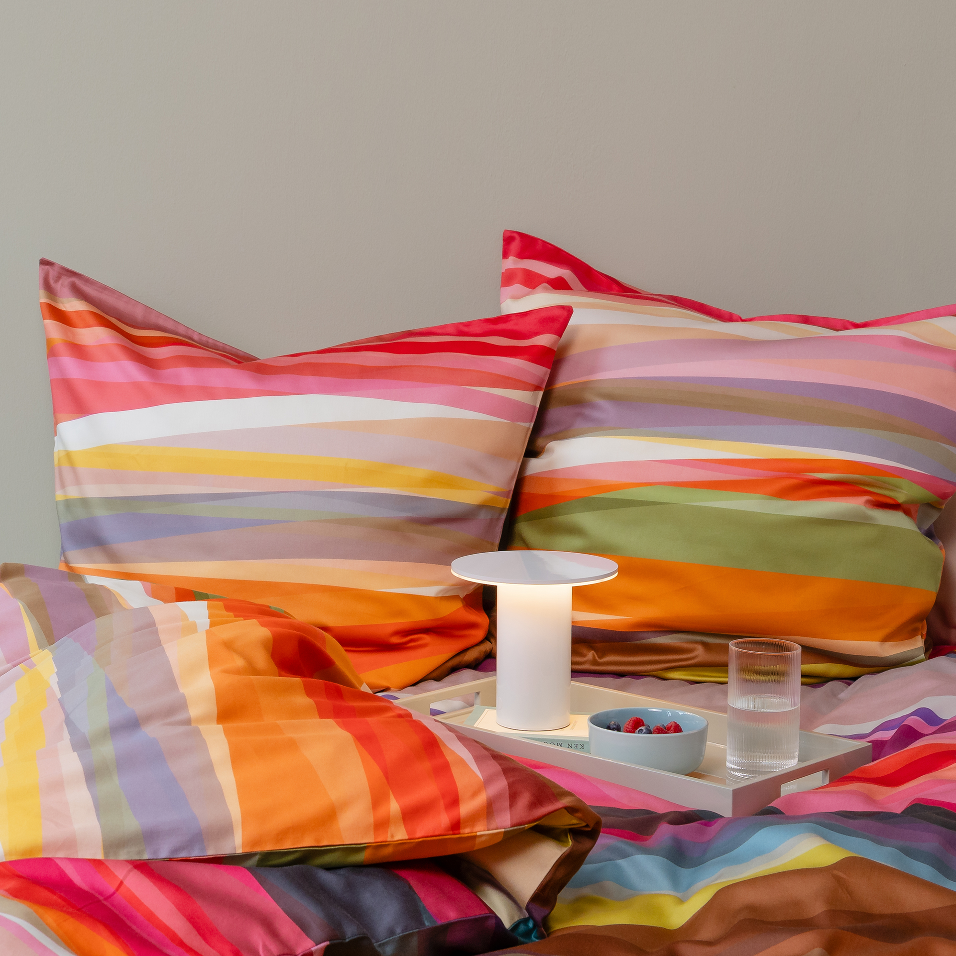 Detailed view of 'Mateo' bed linen (155x220 cm) with colourful stripes on the pillow and duvet. A bedside table with a lamp, berries and a glass of water stands on the bed.
