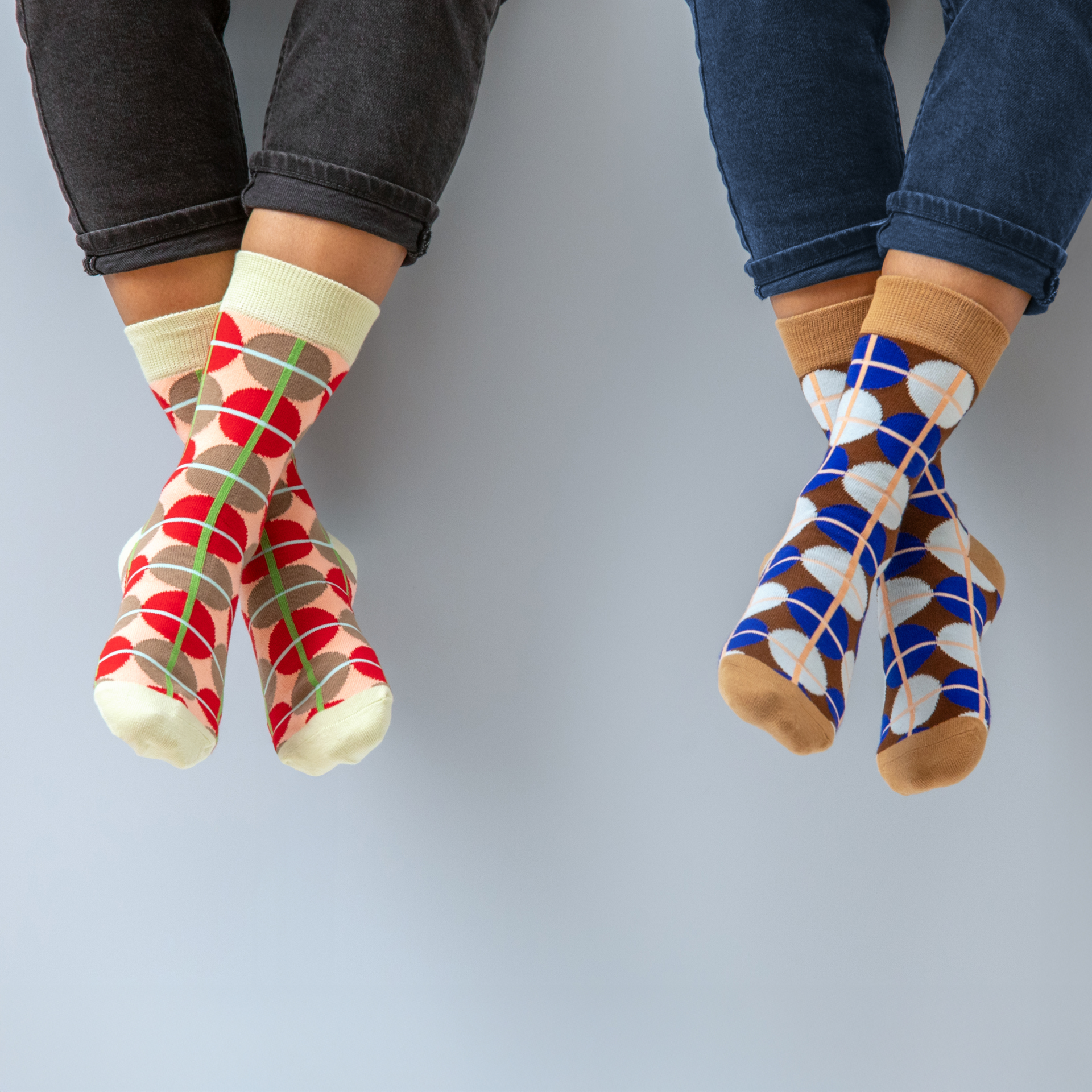 Dos pares de piernas cruzadas con calcetines de diferentes estampados y colores, uno en rojo/marrón/verde y otro en azul/marrón/blanco, a juego con el color de los vaqueros.