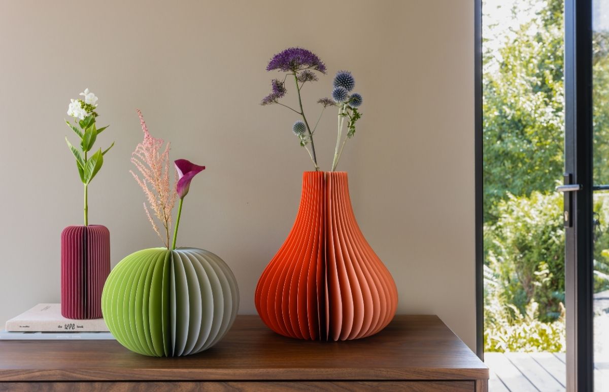 Drei gefaltete Papier-Vasen in verschiedenen Farben und Formen auf einer Holzkommode. Links magentafarbene Zylindervase mit weißer Blume, mittig grüne Kugelvase mit rosa/dunklen Blüten, rechts orange Tropfenvase mit blauen Disteln. Fenster mit Grün.