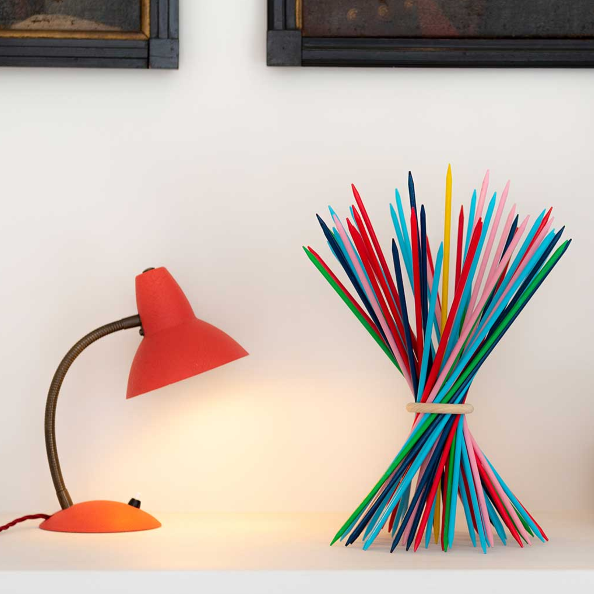 Decoratieve, kleurrijke houten stokjes, samengebonden met een houten ring, naast een rode lamp op een plank voor een witte muur.