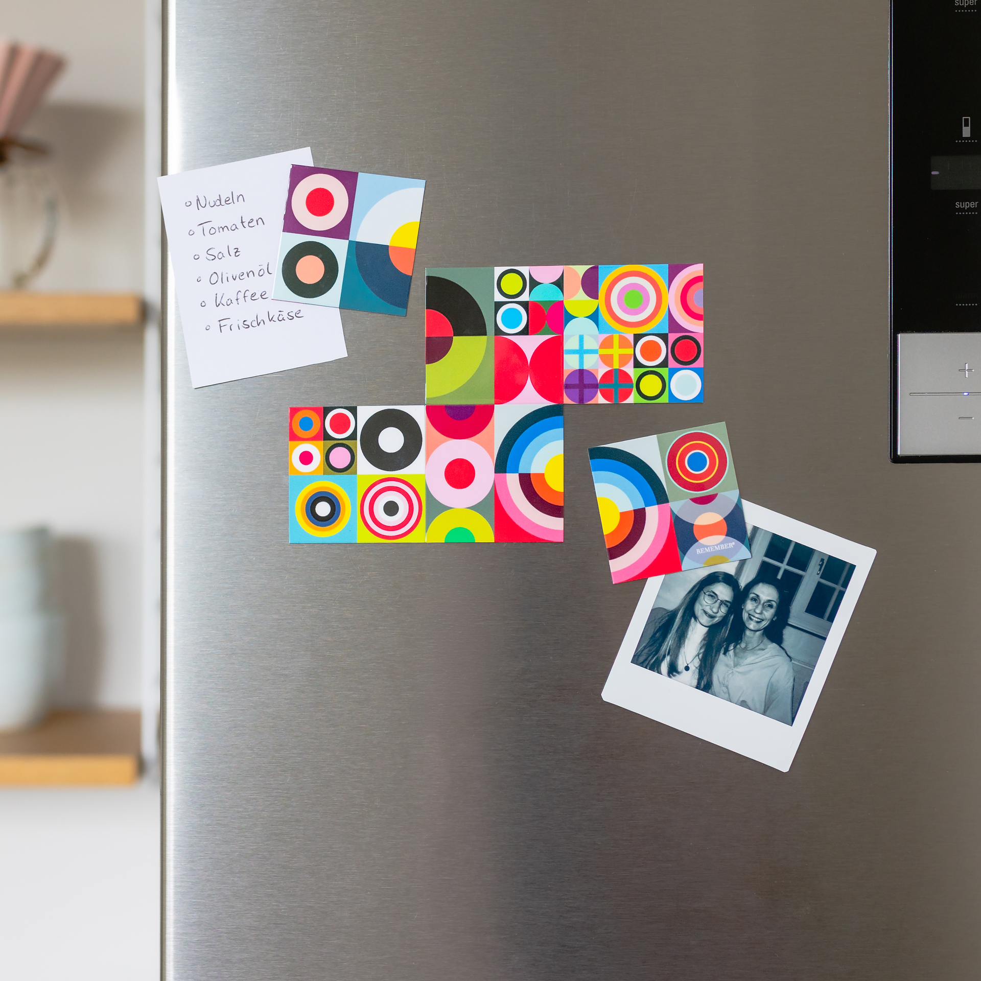A brushed stainless-steel fridge door is adorned with colourful, geometric magnets featuring circular and semi-circular patterns, forming an abstract mosaic of colour. A shopping list and a photo are stuck between them.