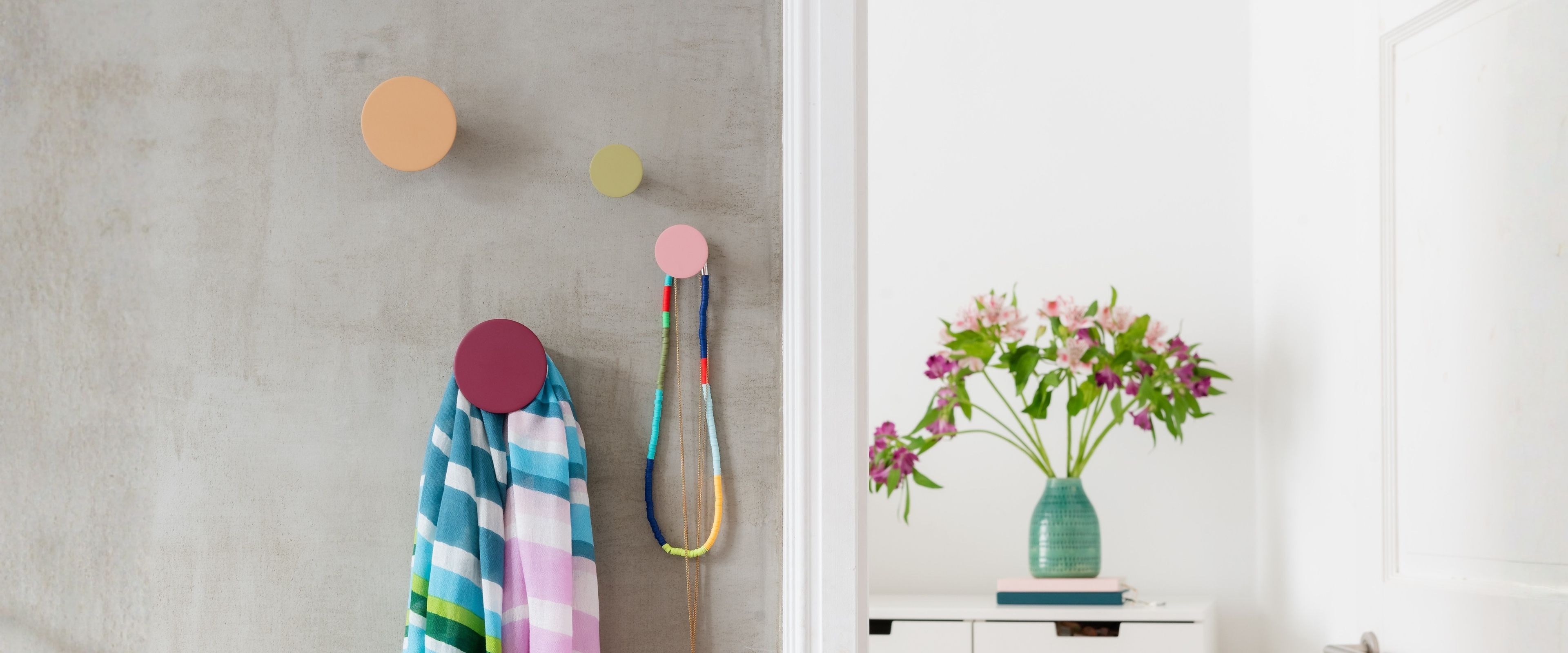 Drei bunte, runde Wandhaken in Pastellfarben an einer grauen Betonwand. Der untere magentafarbene Haken hält einen gestreiften Schal und zwei Perlenketten. Rechts, durch einen Türrahmen getrennt, steht eine Vase mit Blumen.