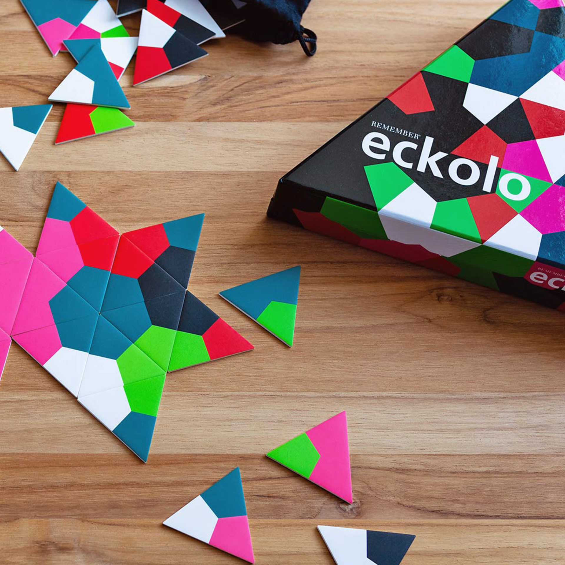 Gioco di posa Eckolo: carte triangolari colorate su una base di legno, parzialmente disposte a formare una stella. Scatola di gioco quadrata sulla destra, con la scritta "eckolo" ben visibile.
