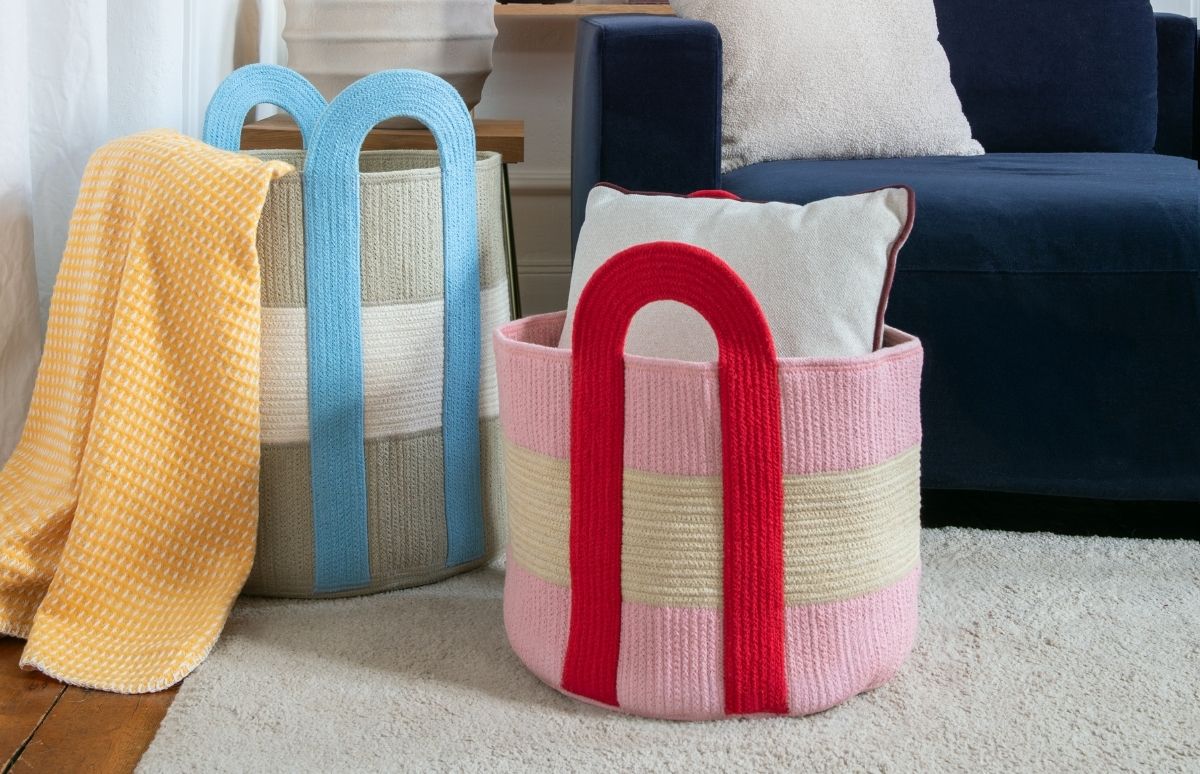 Zwei geflochtene Aufbewahrungskörbe stehen auf einem hellen Teppich. Links: grau-weiß gestreift mit blauen Griffen, mit gelber Waffeldecke. Rechts: rosa-beige gestreift mit roten Griffen, mit hellem Kissen. Dahinter ein dunkelblauer Sessel.