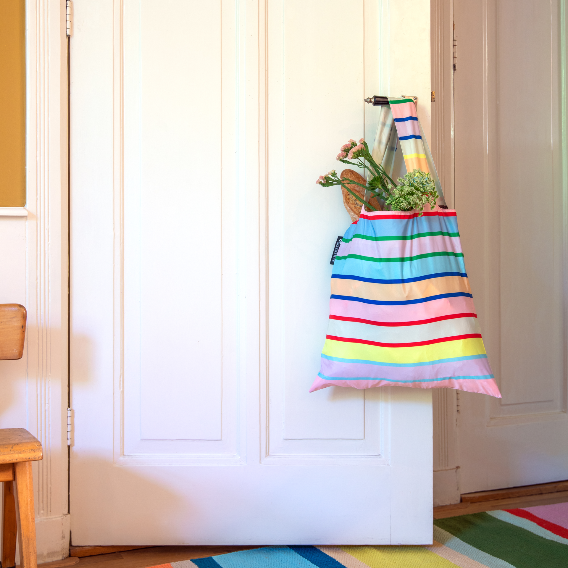 Bunte, faltbare Tragetasche mit horizontalen Regenbogenstreifen in Pink, Blau, Grün und Gelb hängt am Türgriff einer weißen Tür. Gefüllt mit Baguette und frischen Blumen. Auf dem Boden: gestreifter Teppich. Links: Holzstuhl.
