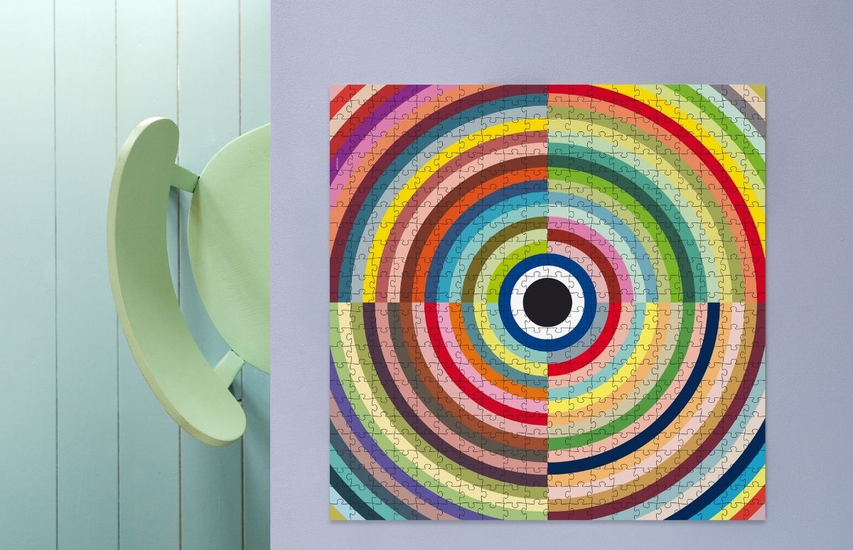 Rundes, farbiges Puzzle mit konzentrischen Regenbogenringen an hellvioletter Wand; Zentrum schwarz. Links ein Stück hellgrüne Stuhllehne vor hellblauer Wand.