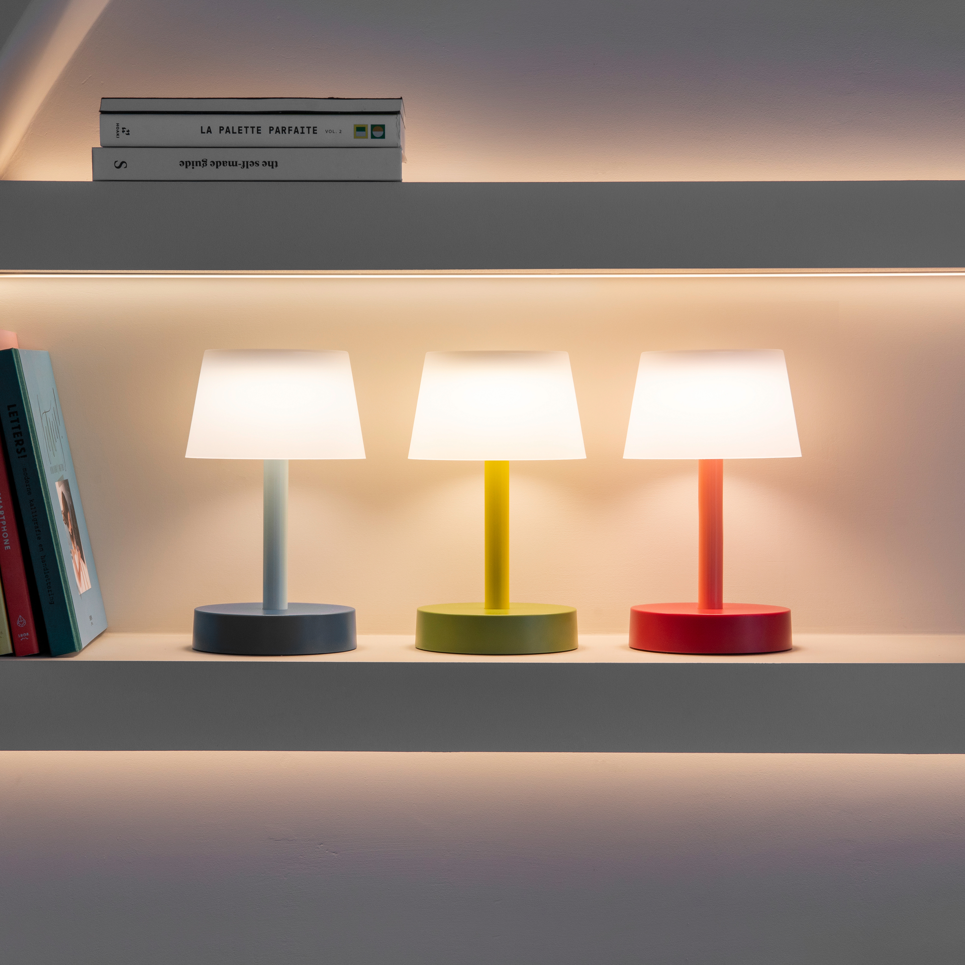 Drie gekleurde Fritz tafellampen op een plank. De lampen in blauw, geel en rood zijn aan en geven warm licht. Boeken liggen ernaast en erboven.