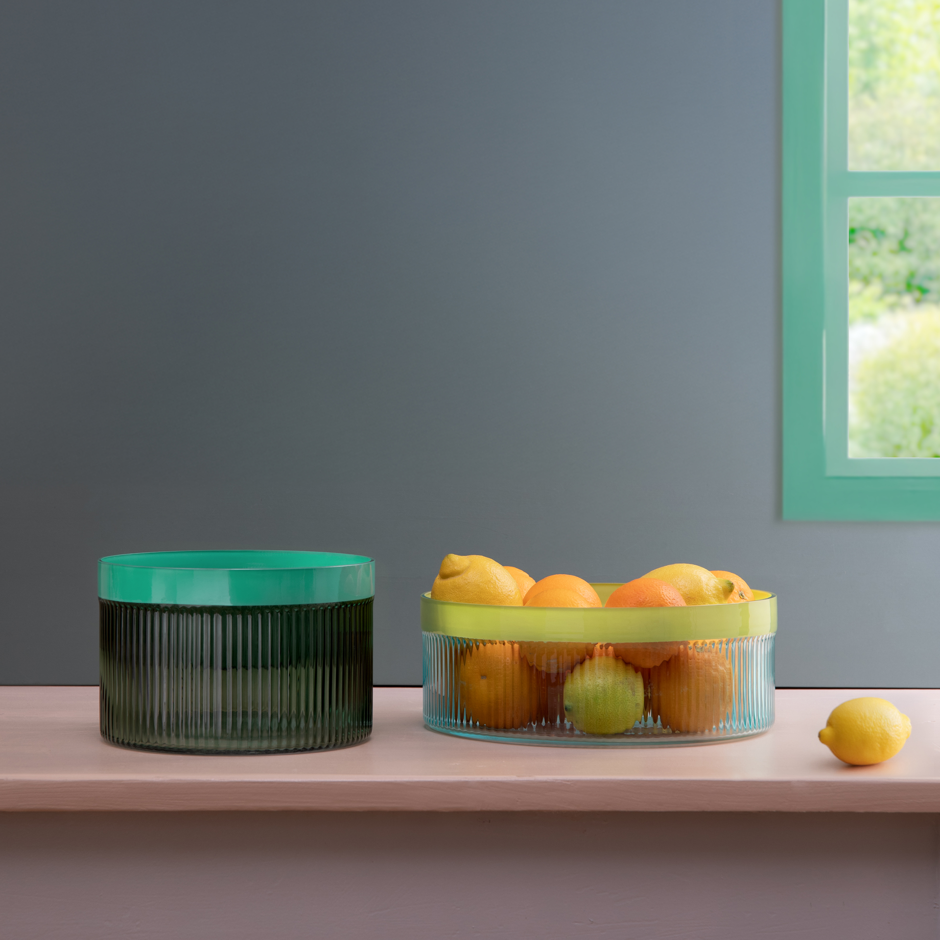 Een groene glazen kom en een 'blauwe' glazen kom gevuld met citrusvruchten op een plank voor een muur en een raam.