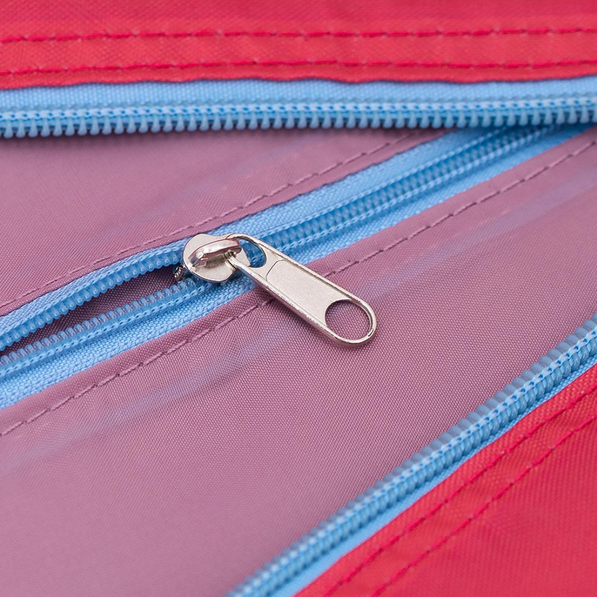 Close-up of the 'Ida' travel bag with zip. The zip is made of metal and lies on pink-coloured fabric. The fabric is surrounded by a light blue zip and red fabric.