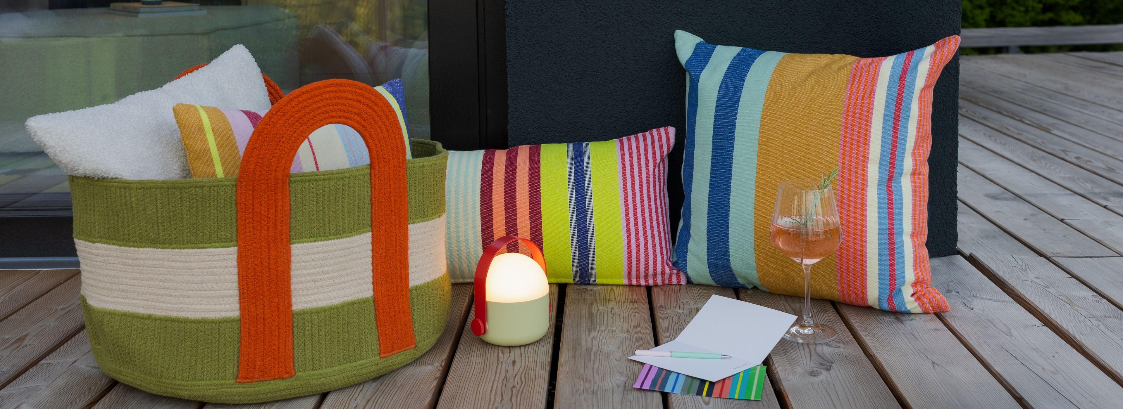Grüner Flechtkorb mit orangen Griffen und weißem Streifen, gefüllt mit weißen Handtüchern und bunten Kissen, steht auf einer Holzterrasse. Daneben leuchtet eine tragbare Lampe. Rechts zwei Kissen, ein Glas Rosé mit Rosmarin, Karte und Stift.