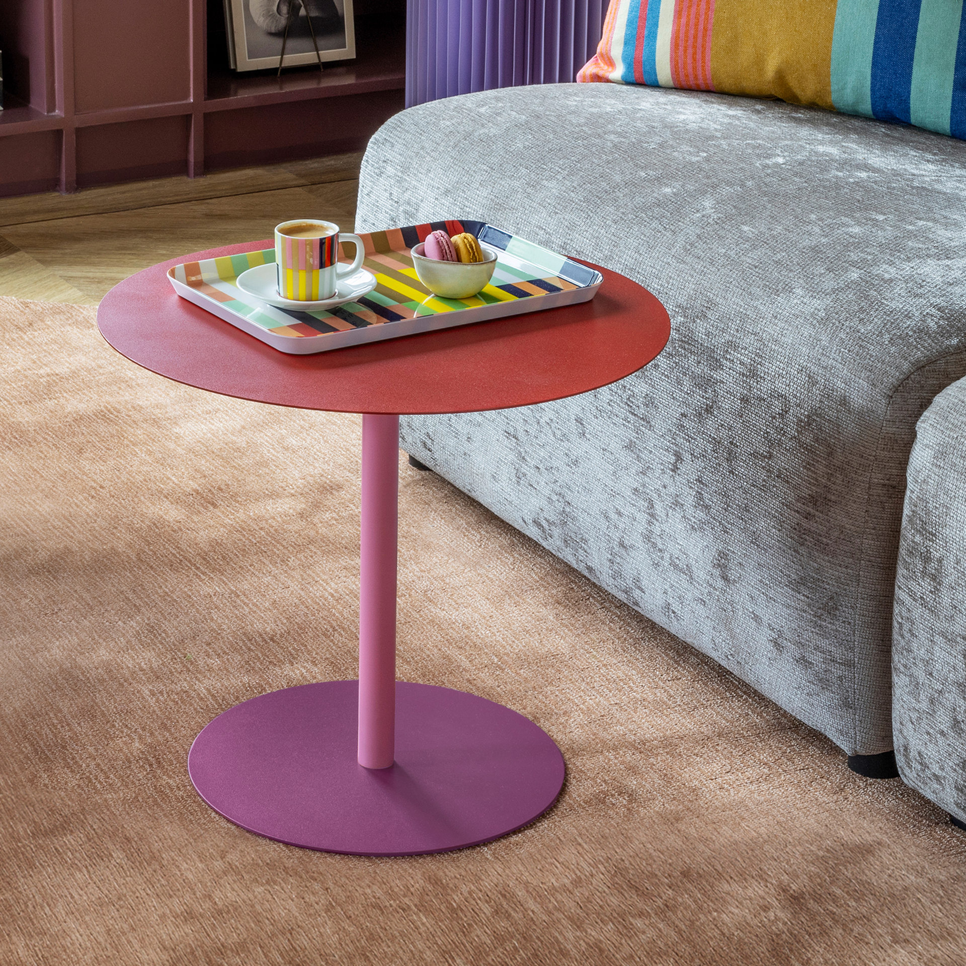 Ronde bijzettafel met een driedelig kleurconcept: het bovenblad is felrood, de cilindrische voet is lichtroze en de ronde sokkel is diep paars. Moderne tafel van gepoedercoat metaal met gladde, matte oppervlakken op een witte achtergrond.