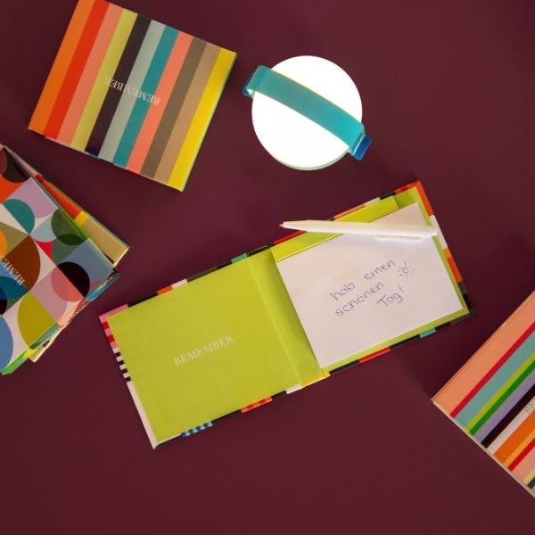 Cadernos coloridos repousam sobre um fundo vermelho escuro. Um livro aberto mostra uma página verde-clara com a frase «REMEMBER » e um bilhete com «Tenha um bom dia!» e um sol. Há uma caneta branca enfiada nele. Um candeeiro redondo com uma fita azul.