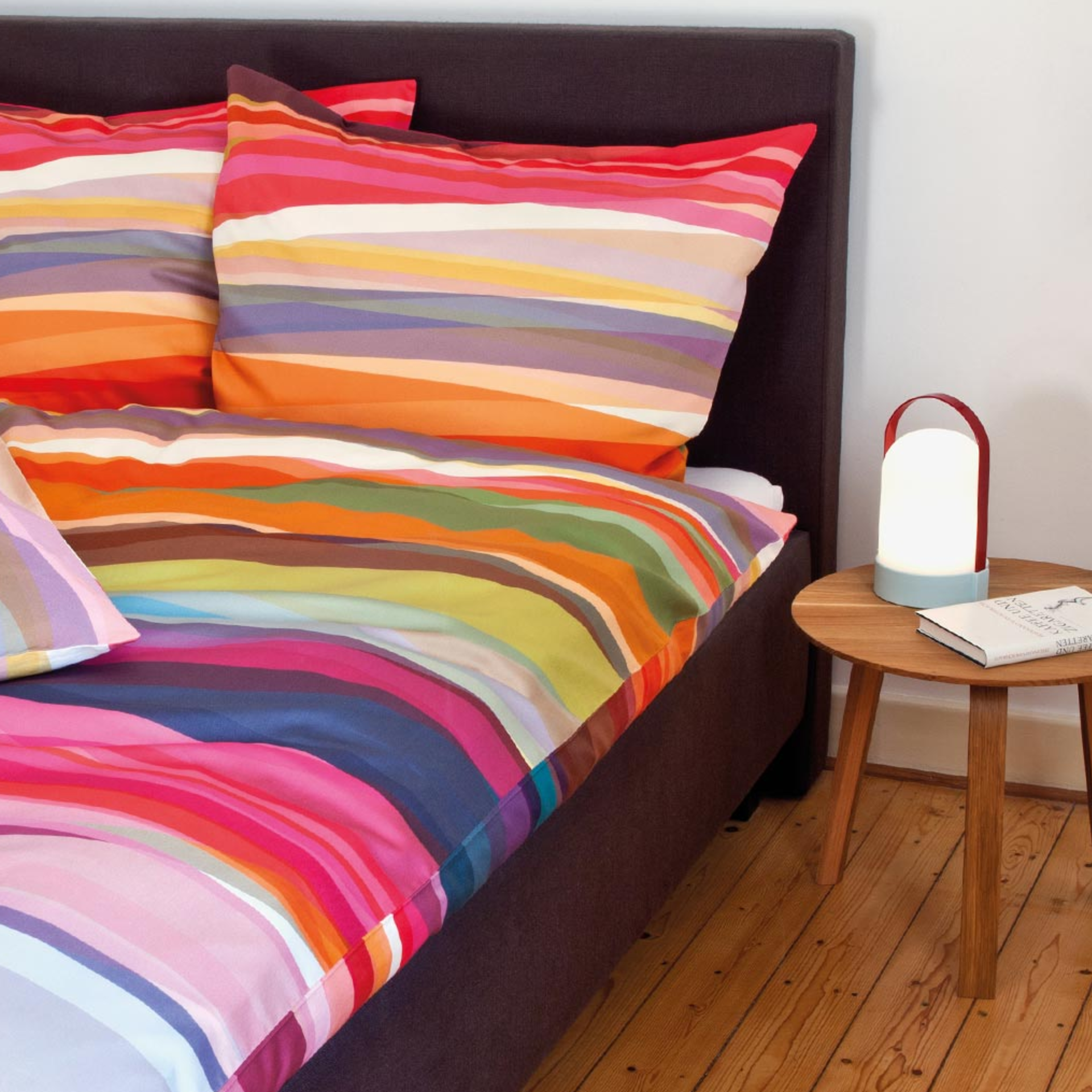 Close-up of a bed with 'Mateo' bed linen in colourful stripes. A bedside table with lamp and book stands next to it on a wooden floor.