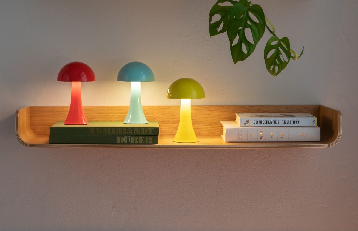 Auf einem hellen Holzregal leuchten drei pilzförmige Tischlampen in Rot, Türkis und Lindgrün und erzeugen ein sanftes Ambiente. Darunter sind Bücher gestapelt, links grüne und rechts weiße. Eine rankende Monstera schmückt oben rechts.