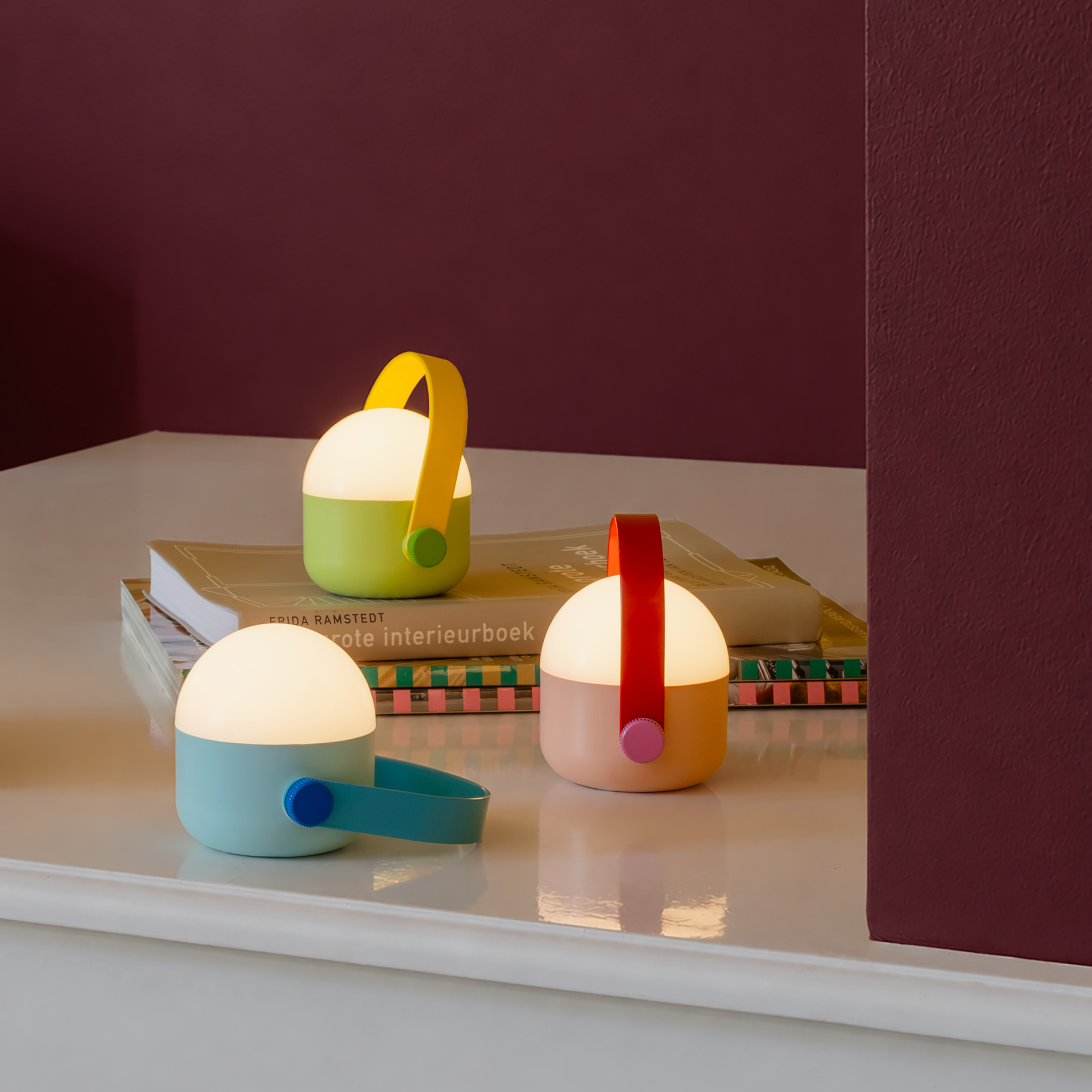 Drei kleine akkubetriebene LED-Leuchten des OLE mini Sets „Remember 30“ leuchten warm auf einer weißen Fläche vor roter Wand. Zu sehen: mintgrün/gelb, hellorange/rot, hellblau/blau. Dimmbar.