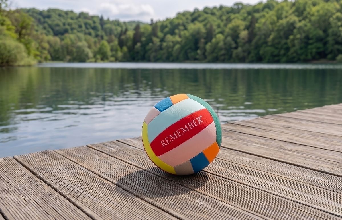 Bunter Volleyball mit roter Aufschrift "REMEMBER" liegt auf sonnenbeschienenem Holzsteg. Der Ball ist in Pastellfarben wie Rot, Blau, Gelb und Grün. Im Hintergrund ein ruhiger See mit dichtem Wald unter leicht bewölktem Himmel.