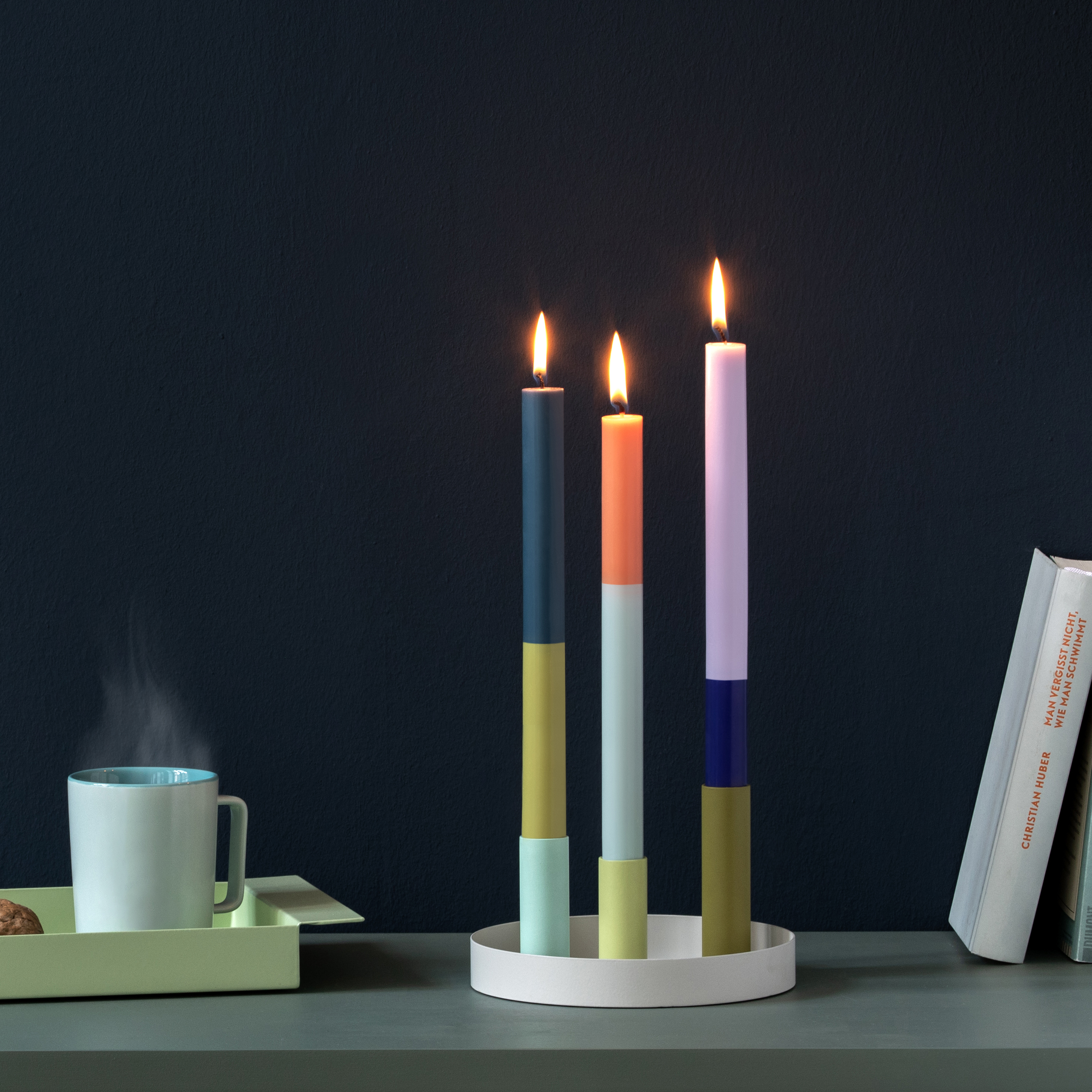 Tres velas de colores encendidas en el portavelas Luna Mini sobre una mesa gris. Junto a ellas, una taza, un libro y una pared oscura.