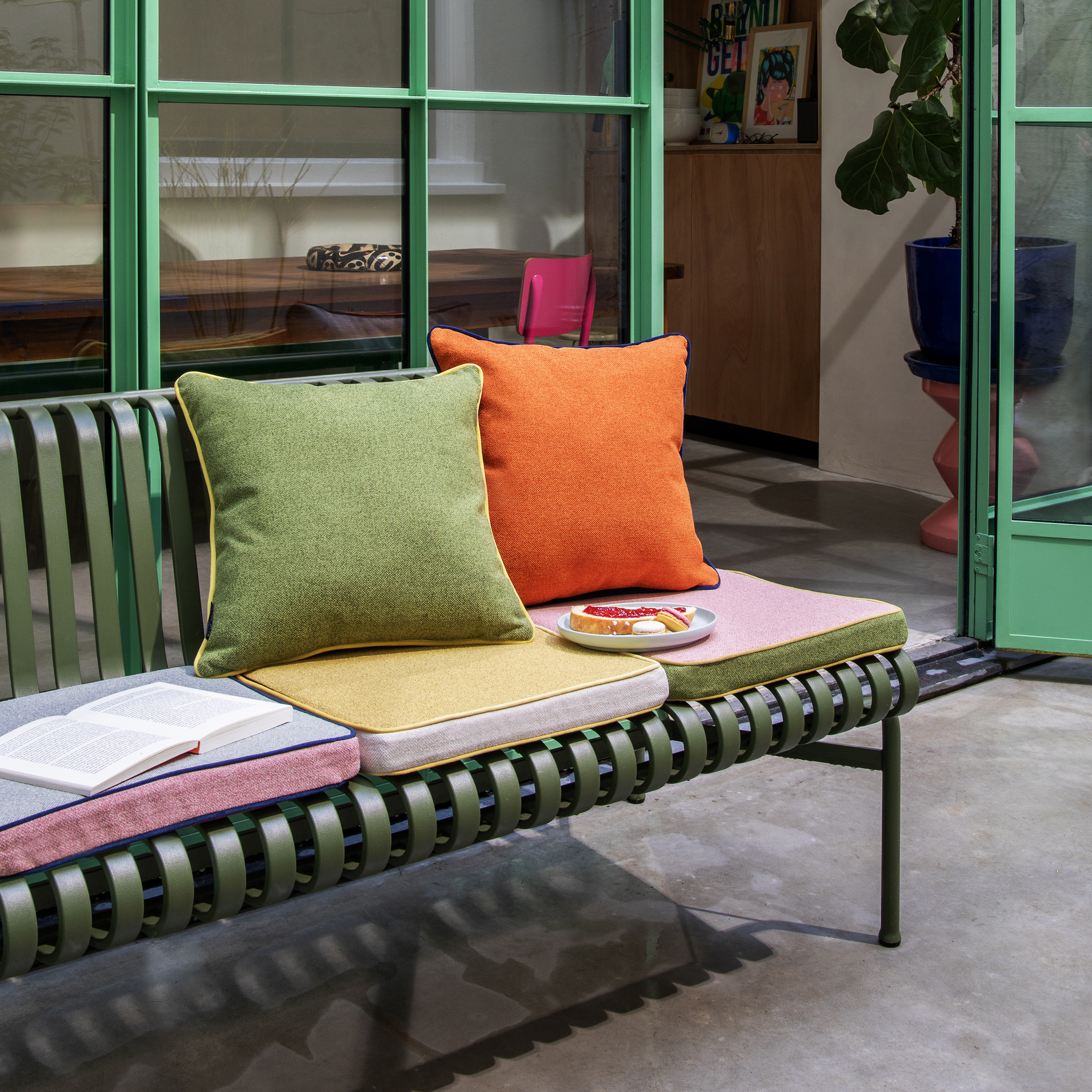 Een groen metalen bankje met zitkussens in roze, geel en grijs. Er liggen een groen en een oranje kussen bovenop. Koekjes en een open boek liggen op een bord.