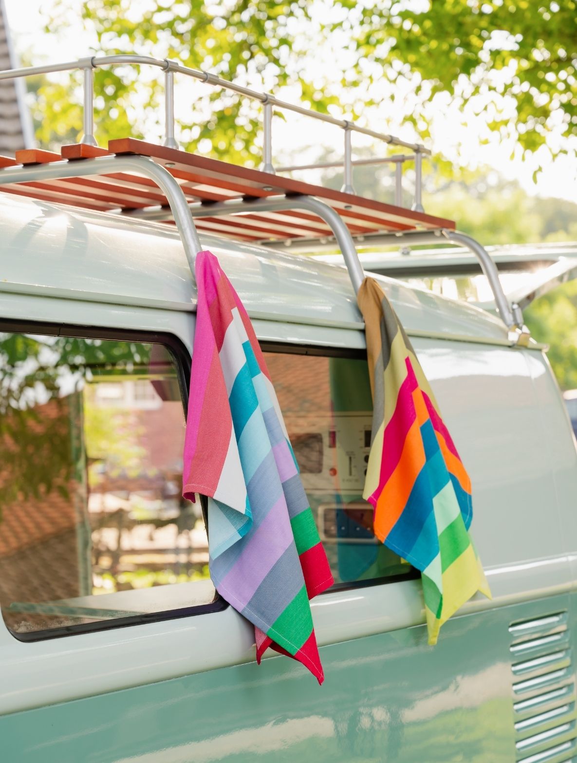 Zwei bunte, gestreifte Handtücher hängen über einem mintgrünen Vintage-Bus am hölzernen Dachgepäckträger. Links Rosa-, Blau- und Grautöne, rechts Gelb-, Orange- und Grüntöne. Hinten unscharfe Bäume und Sonne.