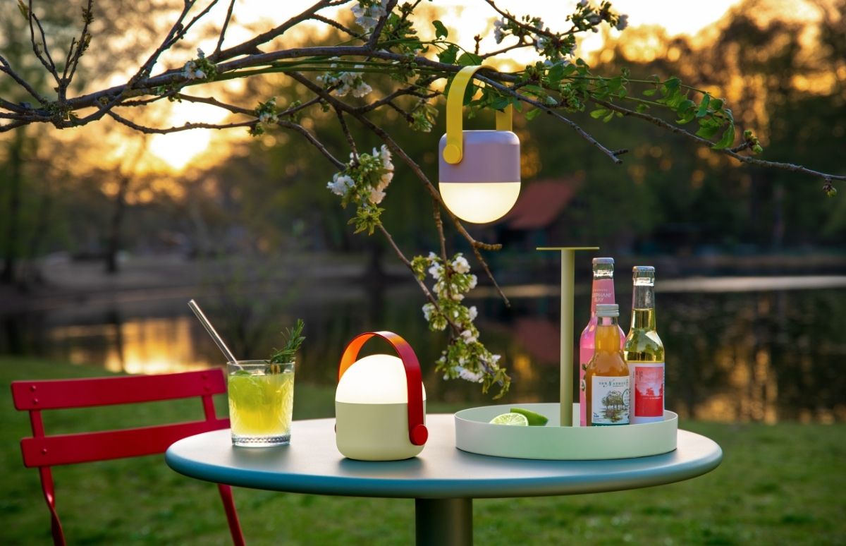 Ein idyllischer Abendtisch im Freien, auf dem ein hellgrünes Getränk mit Rosmarin und zwei leuchtende tragbare Laternen stehen. Eine weitere Laterne hängt von einem blühenden Ast. Im Hintergrund spiegelt sich das goldene Abendlicht auf einem ruhigen See.