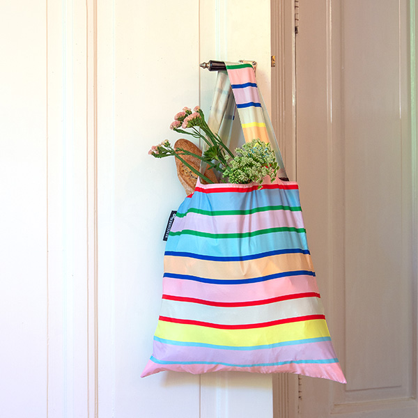 Um saco de compras multicolorido e às riscas está pendurado num puxador de porta. Do saco sobressaem uma baguete estaladiça e flores frescas em tons de rosa e verde-claro. Riscas em tons pastel de rosa, azul, amarelo e verde.