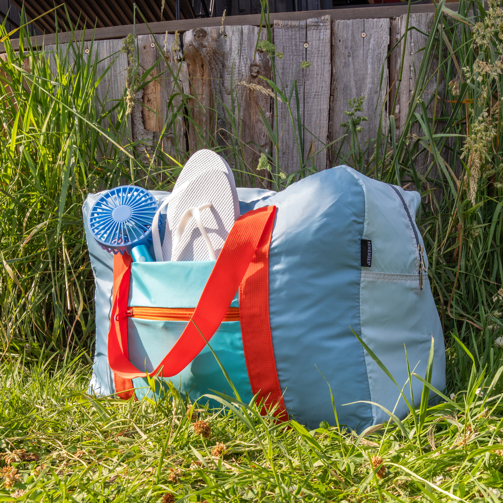 Borsa da viaggio "Tom" azzurra con cinghie arancioni nell'erba. All'interno: infradito bianche e un mini ventilatore. Sullo sfondo una staccionata di legno.
