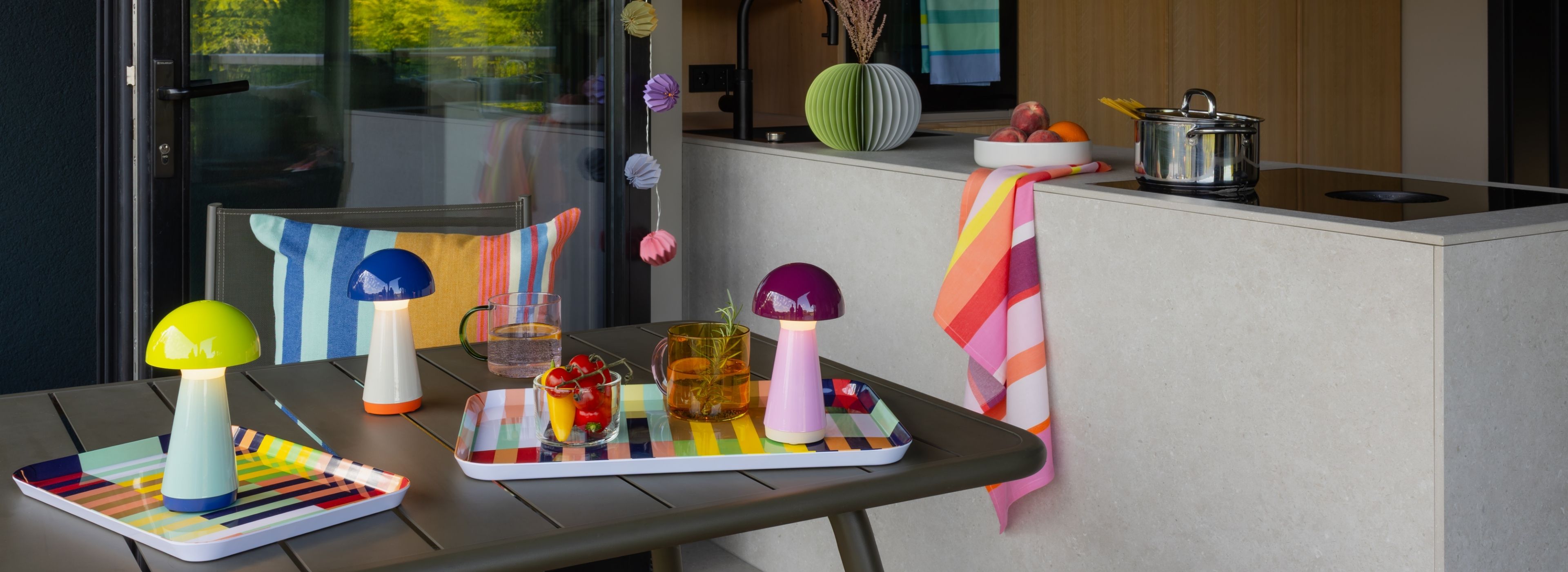 Drei farbenfrohe Pilzlampen stehen auf einem Gartentisch, umgeben von zwei gestreiften Servierplatten mit kühlen Getränken und frischem Snack-Gemüse. Im Hintergrund sind moderne Küchenzeile, Herd, Obstschale und ein buntes Handtuch zu sehen.